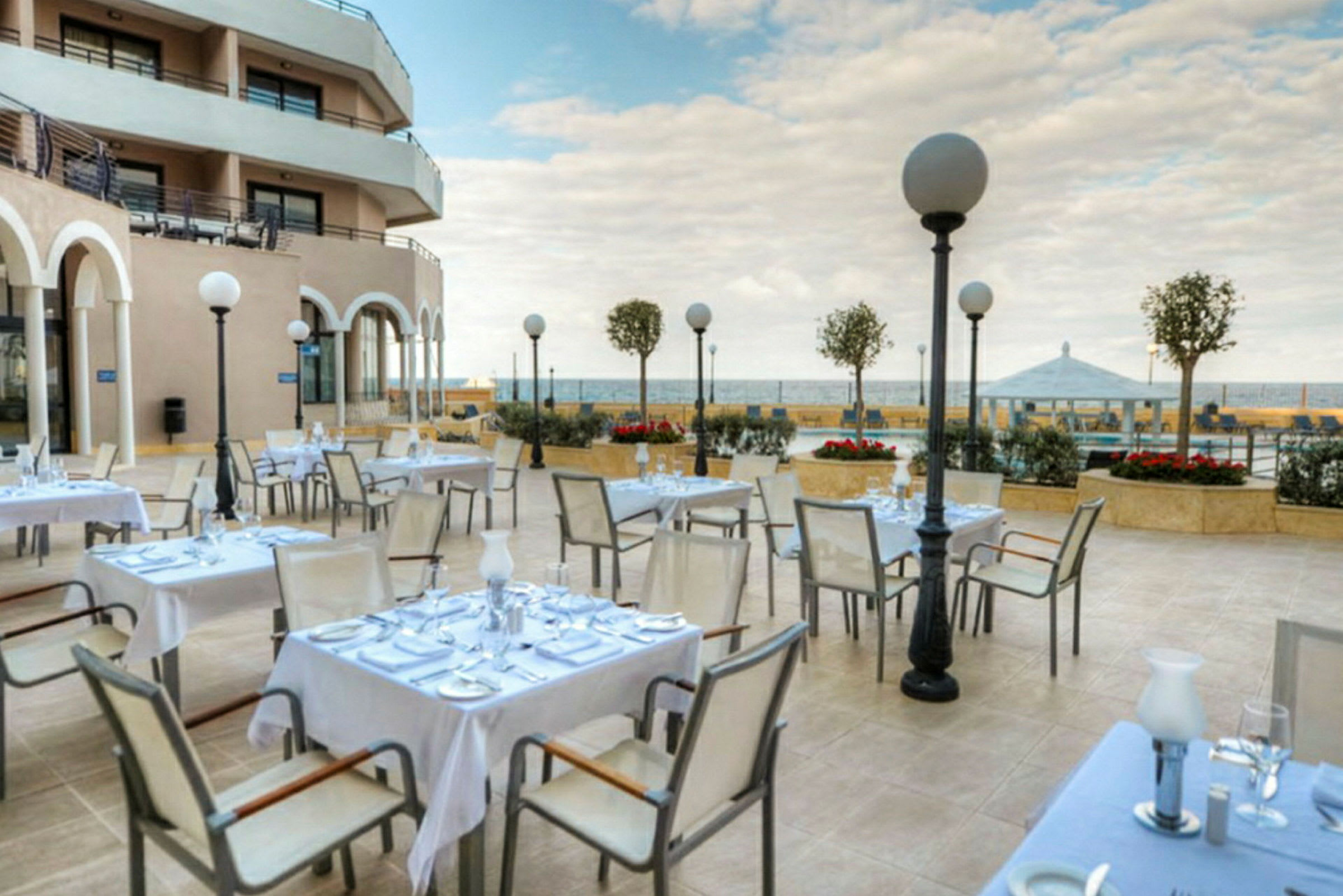 En uteservering vid ett hotell med dukade bord och stolar, omgiven av blomsterkrukor och utsikt över havet. (AI-genererad text)