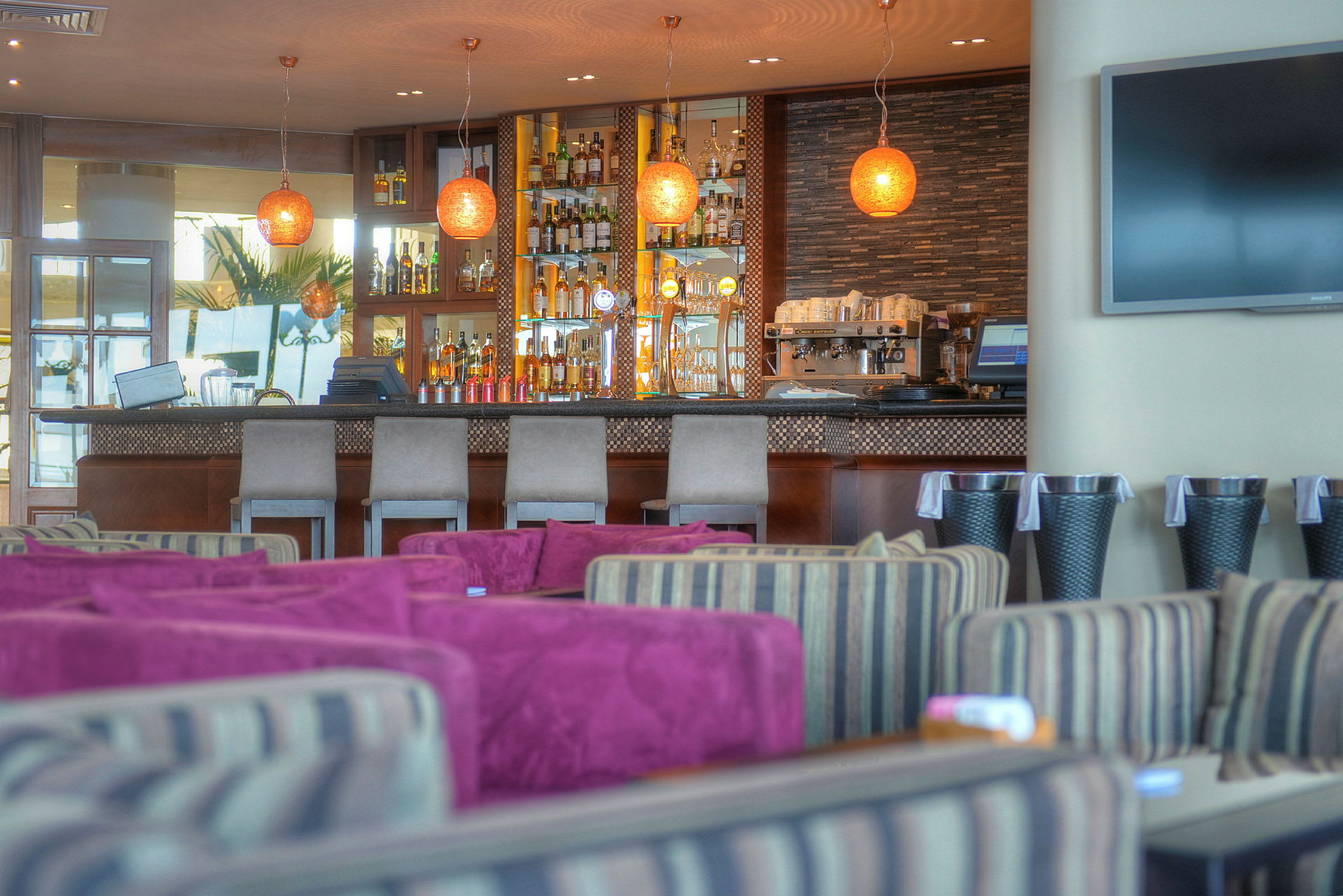En interiörbild av en elegant bar med rader av drycker, prydliga barstolar och moderna lampor. I förgrunden syns sittplatser med färgglada kuddar. (AI-genererad text)