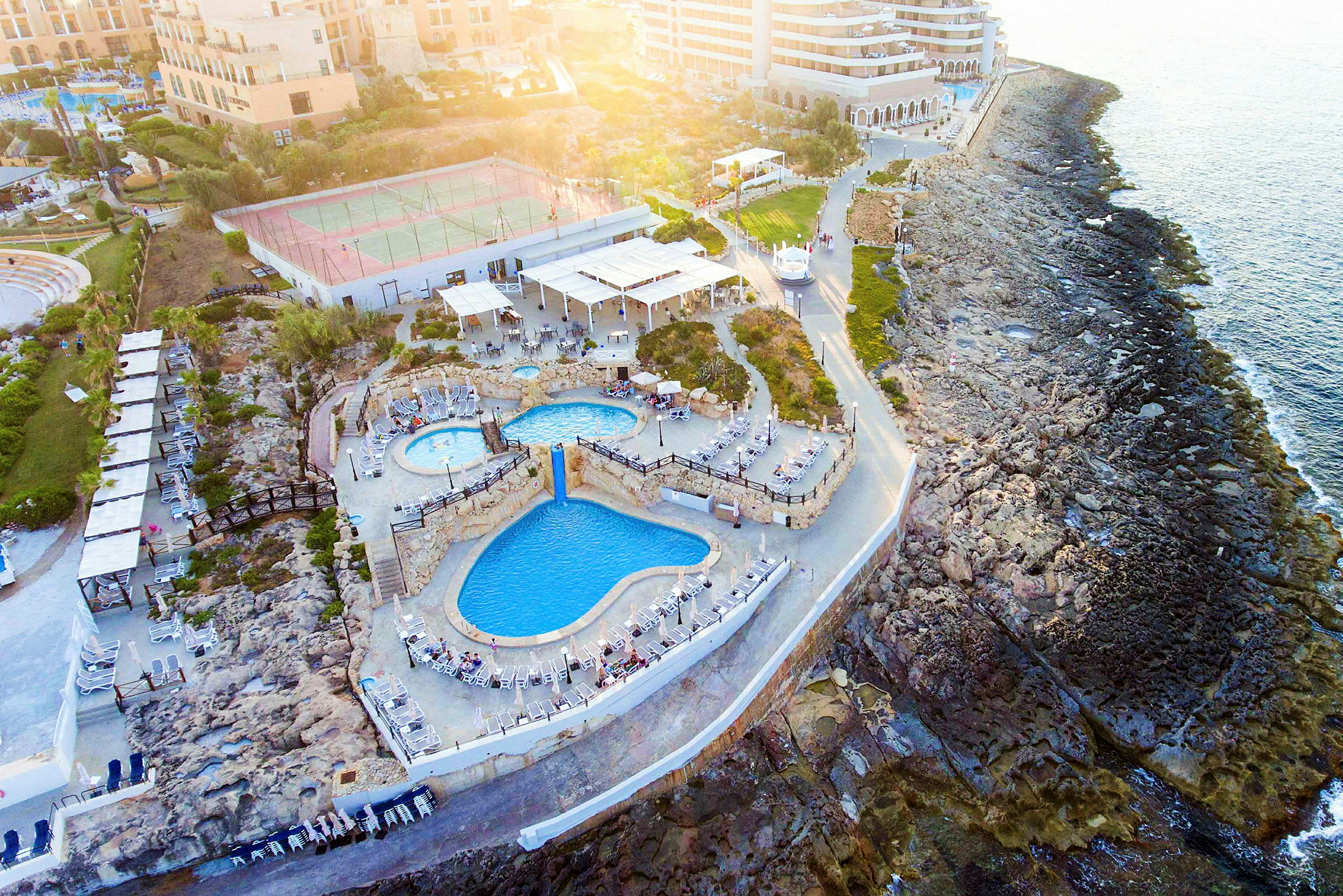 Bilden visar en resort vid kusten med flera pooler och solstolar, omgiven av klippor och byggnader i bakgrunden. (AI-genererad text)