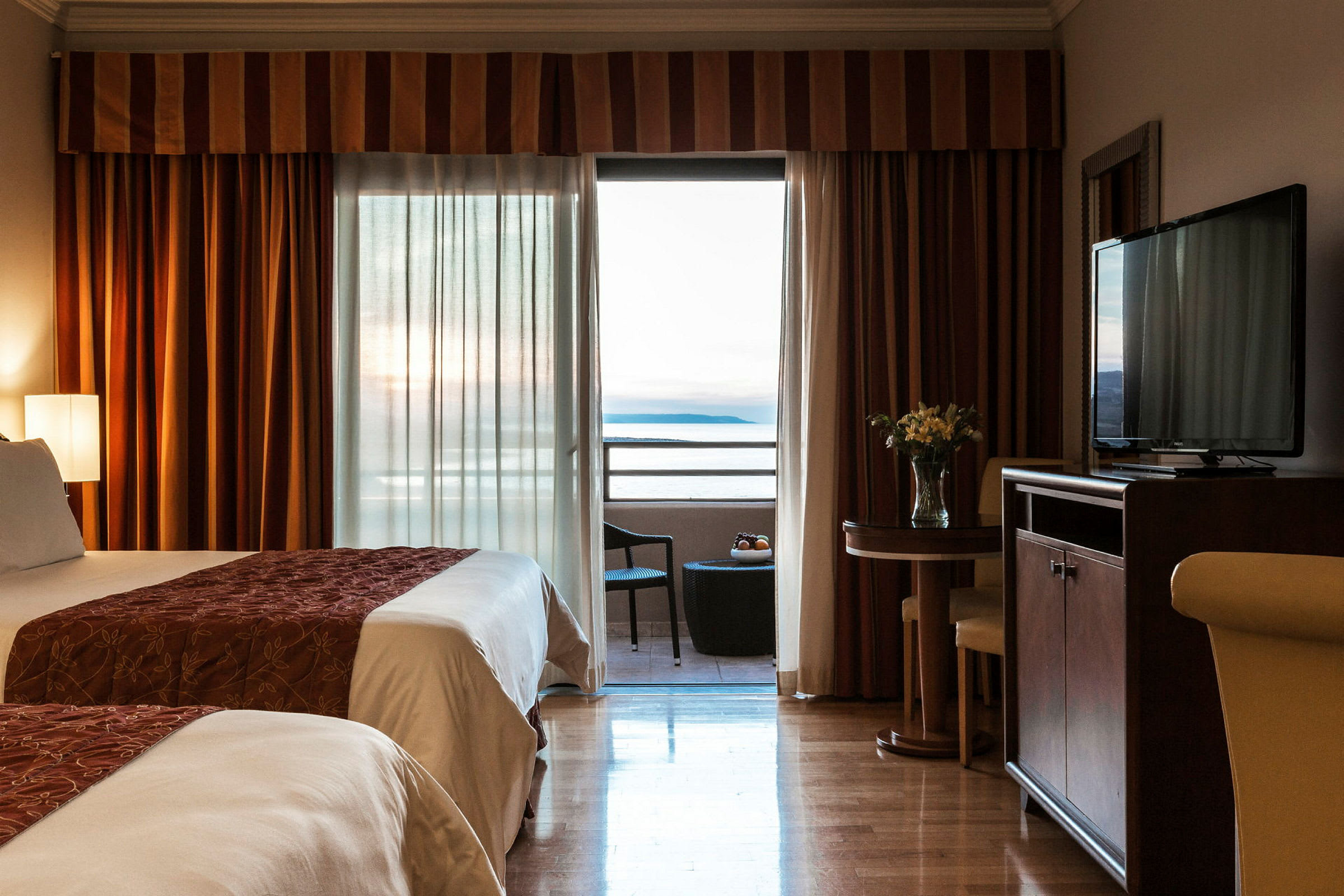 Ett hotellrum med två bäddar, en balkong med utsikt över havet, och ett mindre bord med stolar. (AI-genererad text)