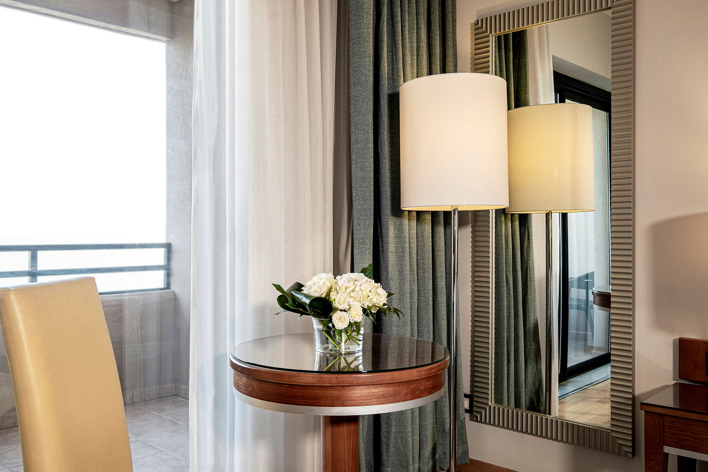 Ett elegant hotellsittområde med en stol, ett litet runt bord med en blomsterbukett, en stor spegel och en golvlampa sett genom ett fönster. (AI-genererad text)