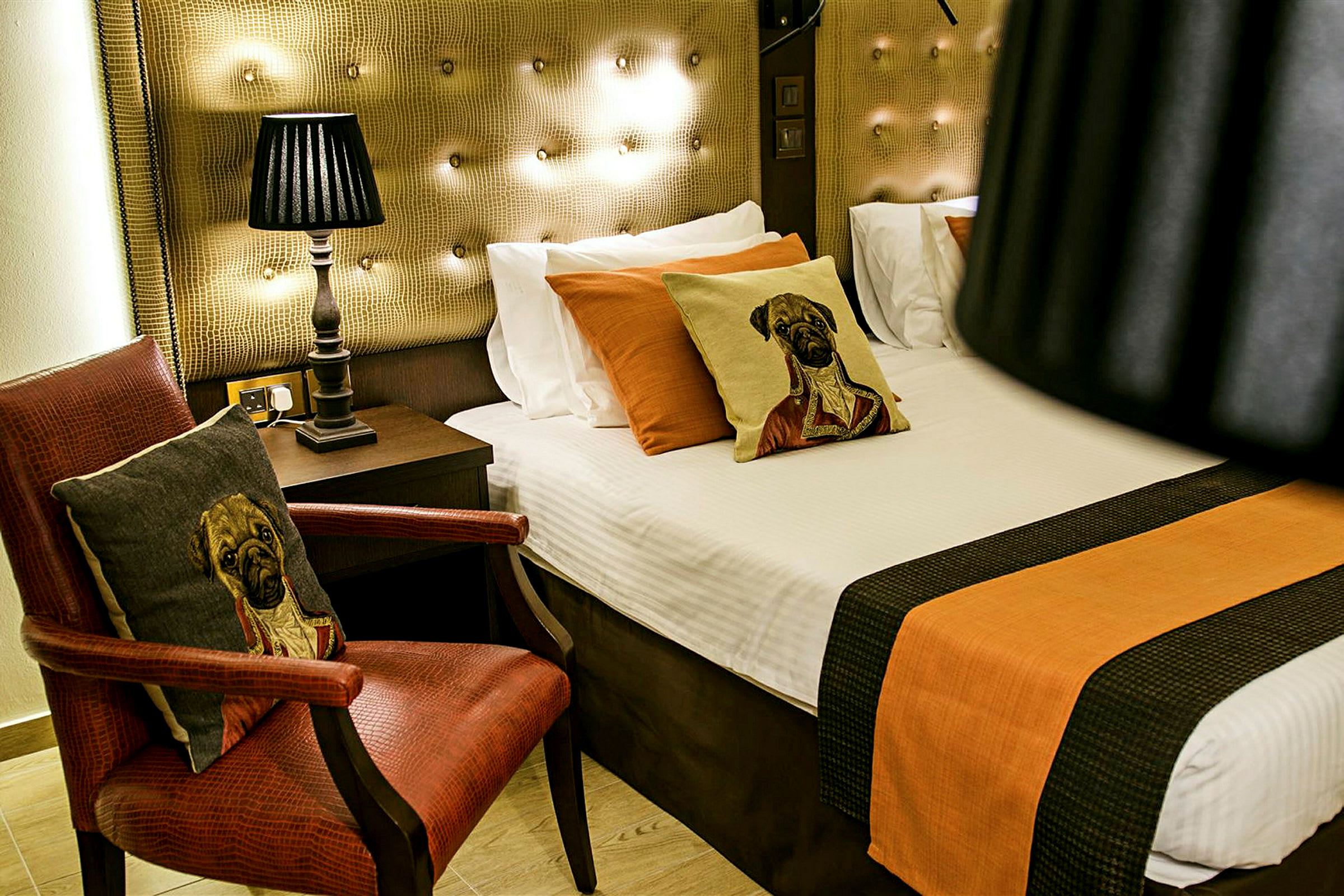 En hotellrumsmiljö med en säng, kuddar med motiv av hundar, en bordslampa och en röd fåtölj med ett matchande kudde. (AI-genererad text)