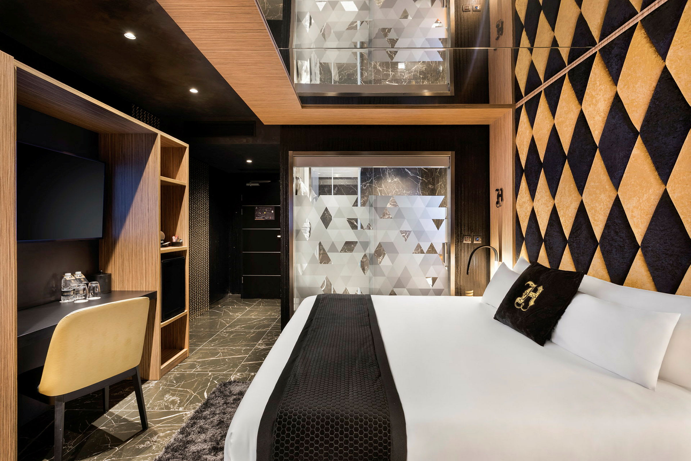 Ett hotellrum med en modern inredning. Rummet har en stor säng, geometriska dekorväggar och ett skrivbord med stol. En frostad glasvägg skiljer badrummet från sovdelen. (AI-genererad text)
