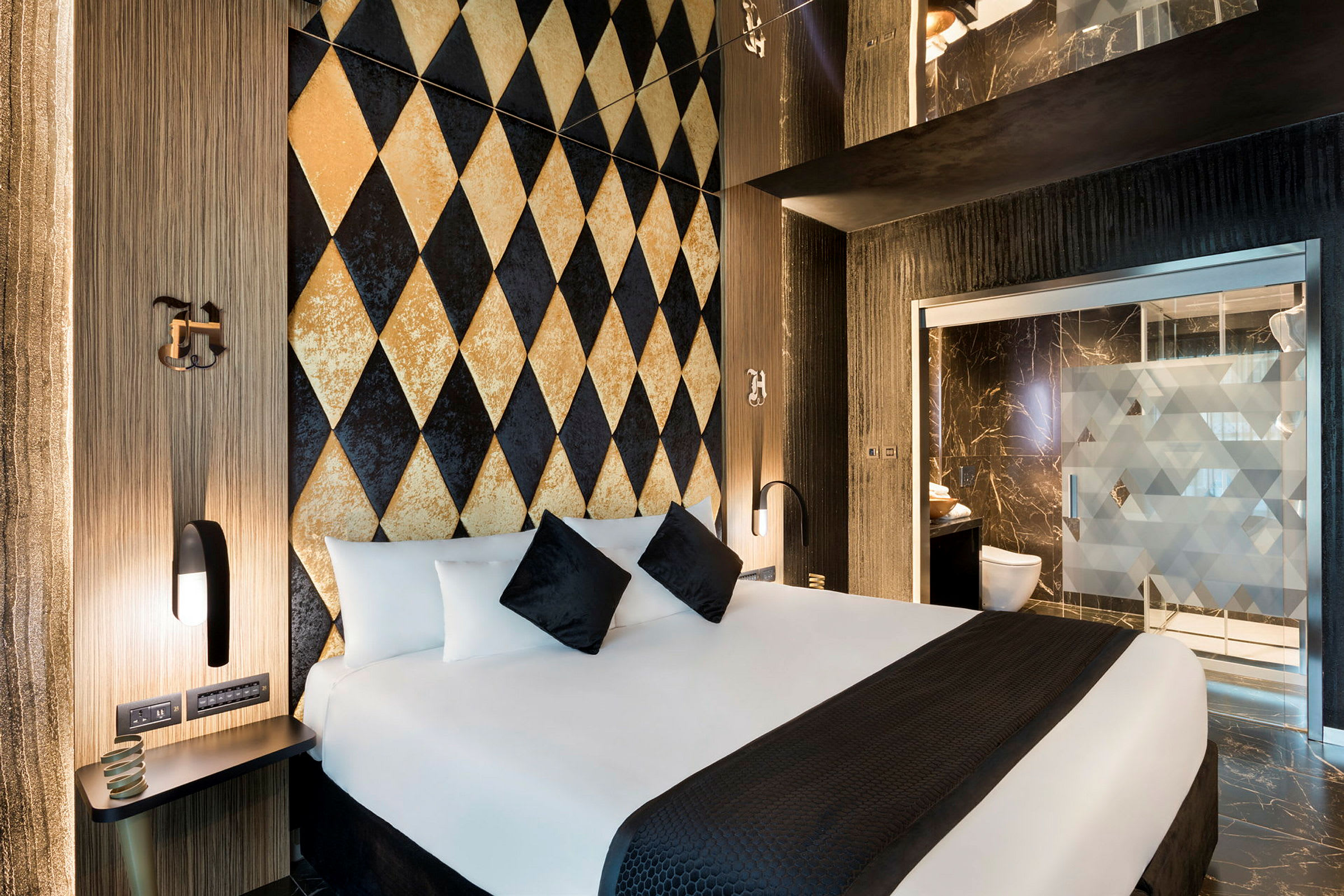 En modern hotellrum med dekorationsvägg i harlekinmönster och en stor säng med vita och svarta kuddar. Badrummet ses i bakgrunden. (AI-genererad text)