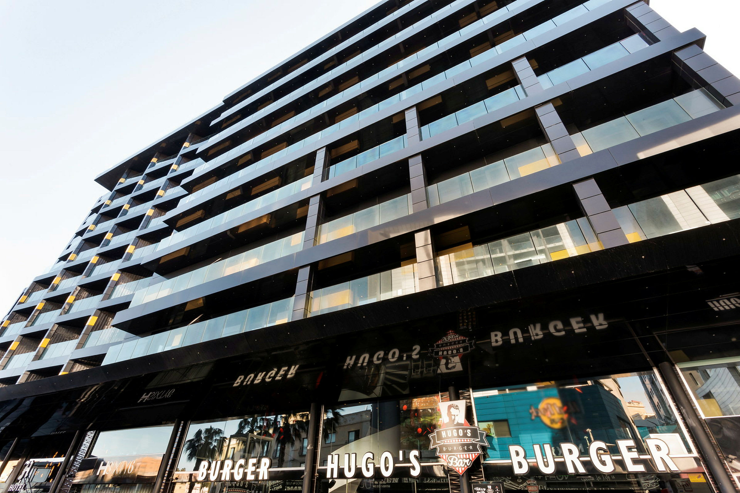 En modern byggnad med glas- och metallfasad samt skyltar för ett hamburgerställe kallat Hugo's Burger. (AI-genererad text)