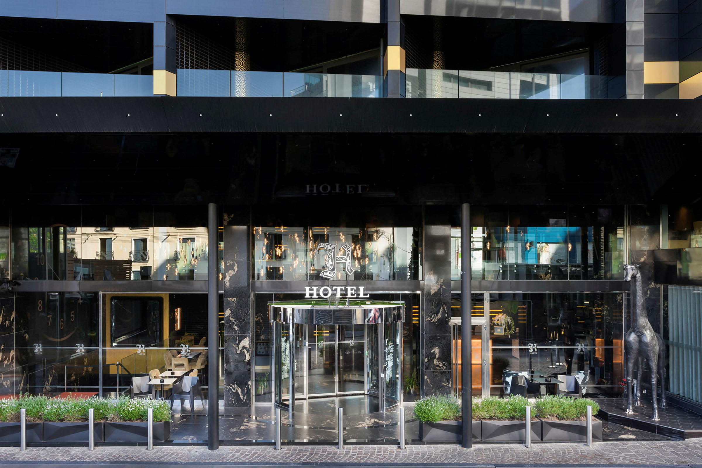 Framsidan av ett hotell med glasdörrar och en dekorativ silverfärgad giraffstaty vid sidan av entrén. (AI-genererad text)