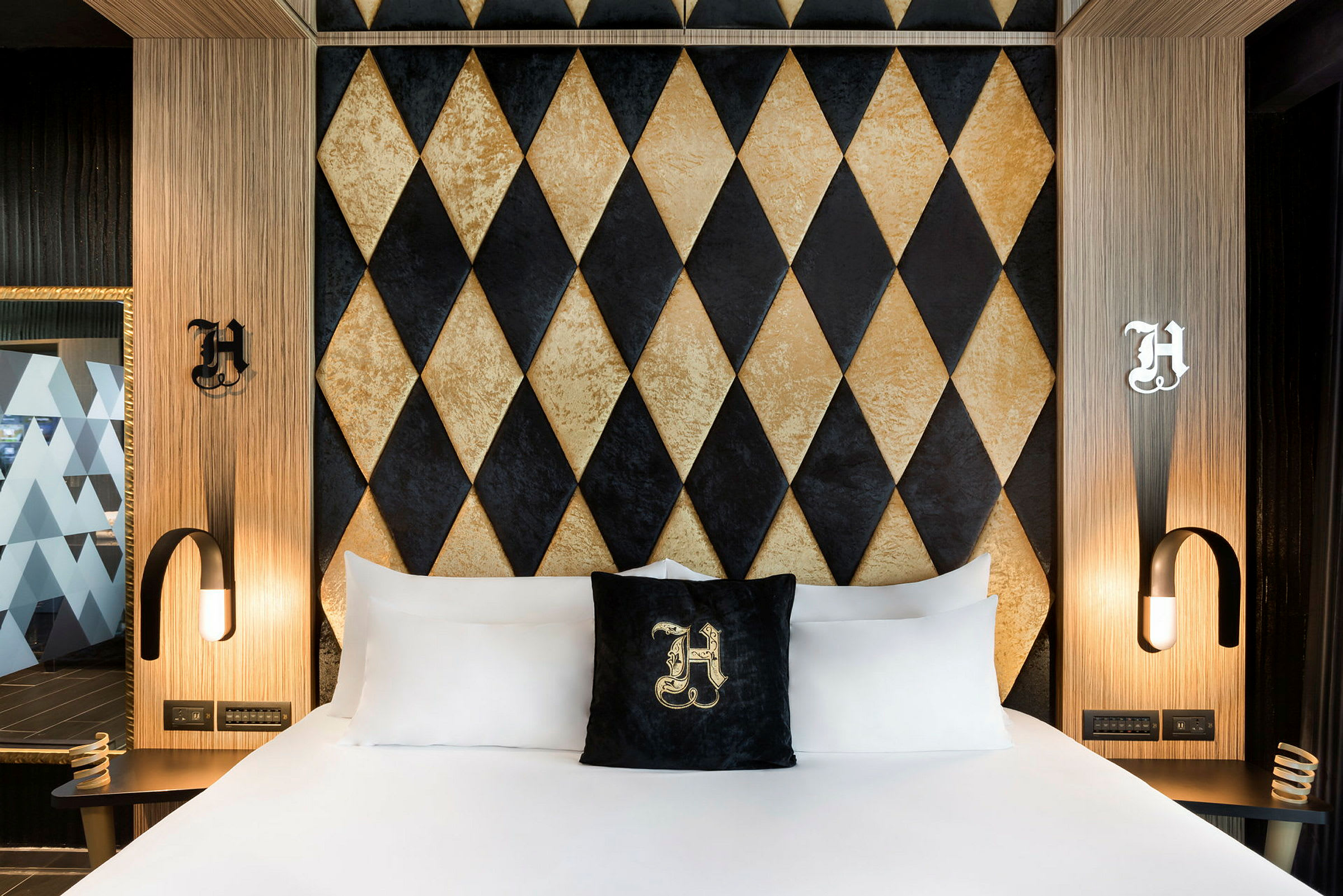 Ett hotellrum med en säng som har vita lakan och en prydnadskudde med en dekorativ bokstav. Bakom sängen finns en vägg med diamantmönster i svart och guld. (AI-genererad text)