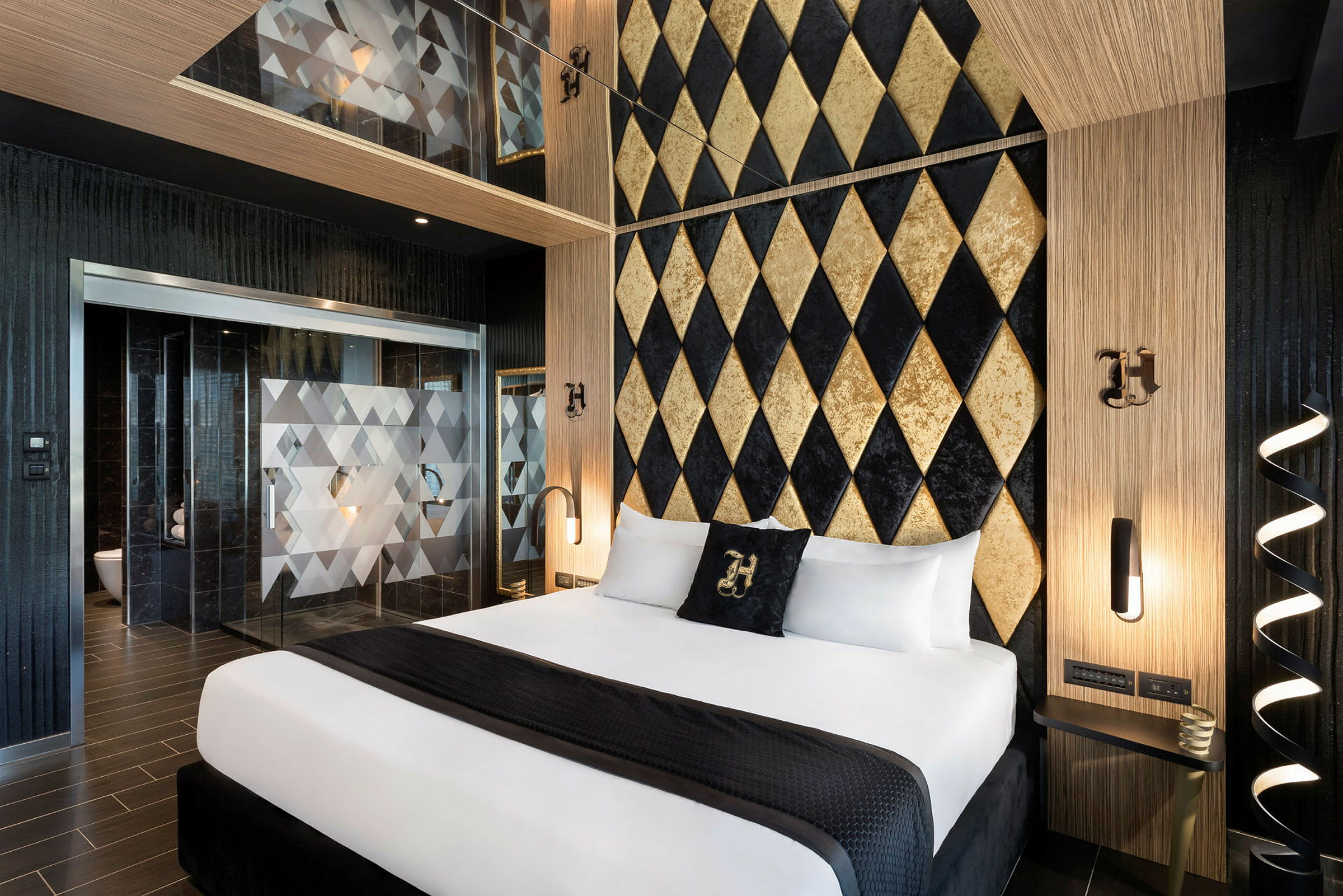 Ett hotellrum med en elegant design. En dubbelsäng med vita lakan och en svart filt är centralt placerad. Bakgrunden är en vägg med guldfärgade och svarta diamantmönster. Det finns separerade glasdörrar med geometriska detaljer som leder till ett badrum. (AI-genererad text)