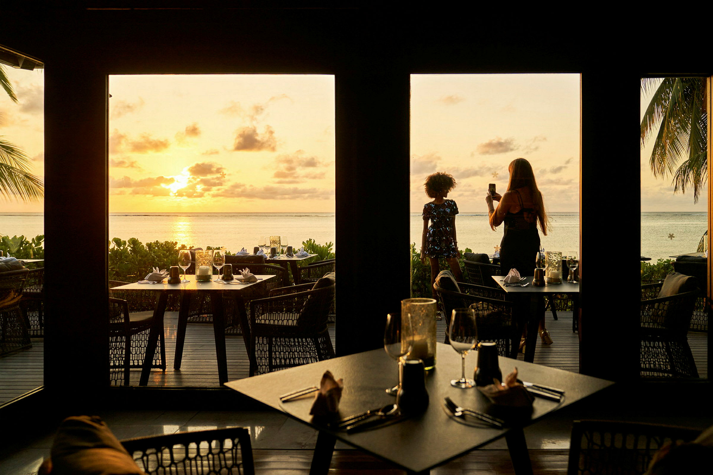 Bilden visar en restaurang med utsikt över en strand vid solnedgången, med bord uppställda både inomhus och utomhus, samt palmer och havet i bakgrunden. (AI-genererad text)