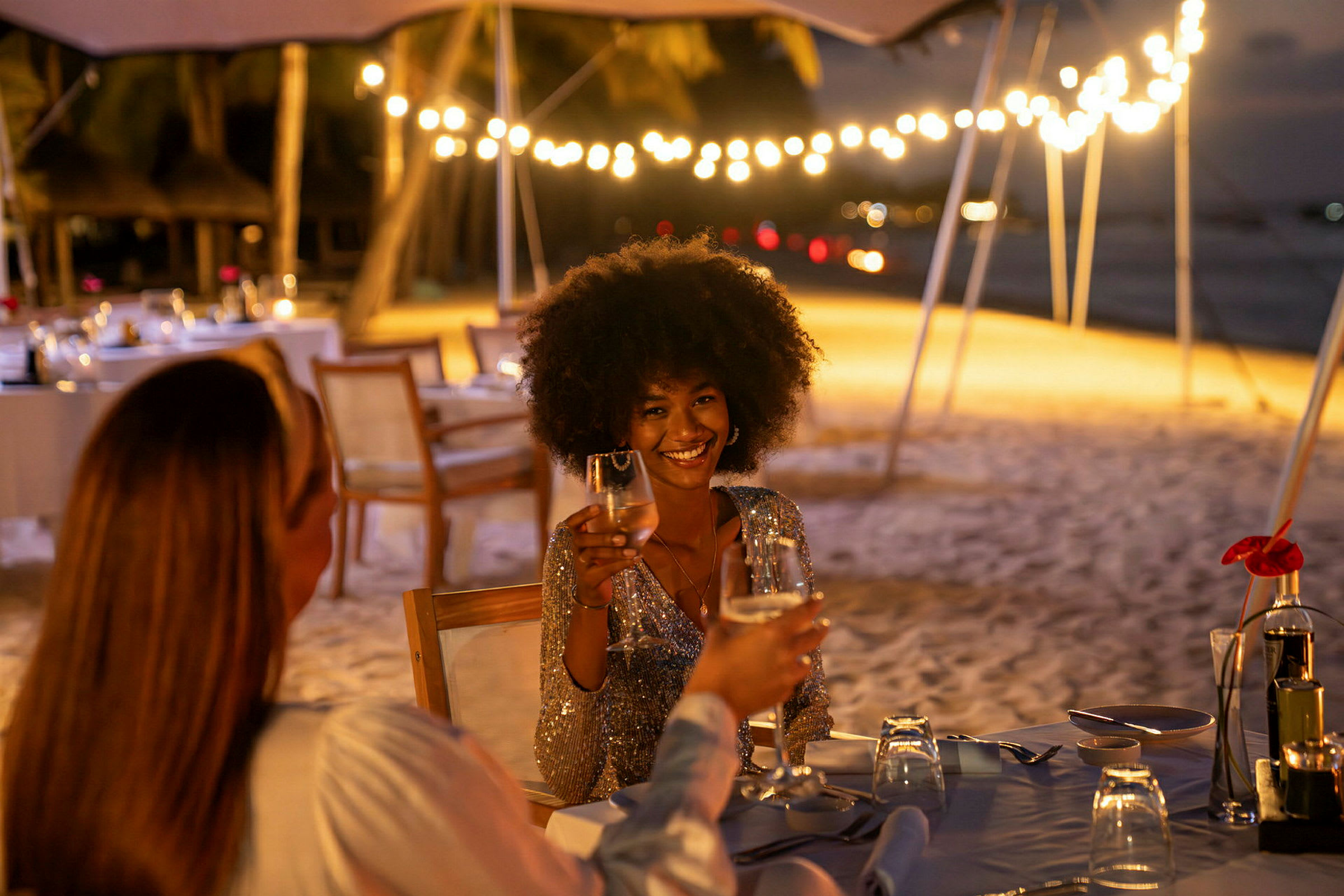 Två personer sitter vid ett dukat bord på en strand med ljusslingor i bakgrunden. (AI-genererad text)