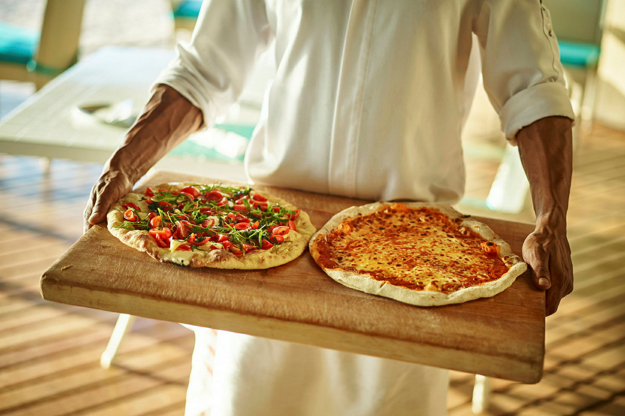 En person bär två pizzor, en med färska tomater och ruccola och en med smält ost, på en träbricka. (AI-genererad text)