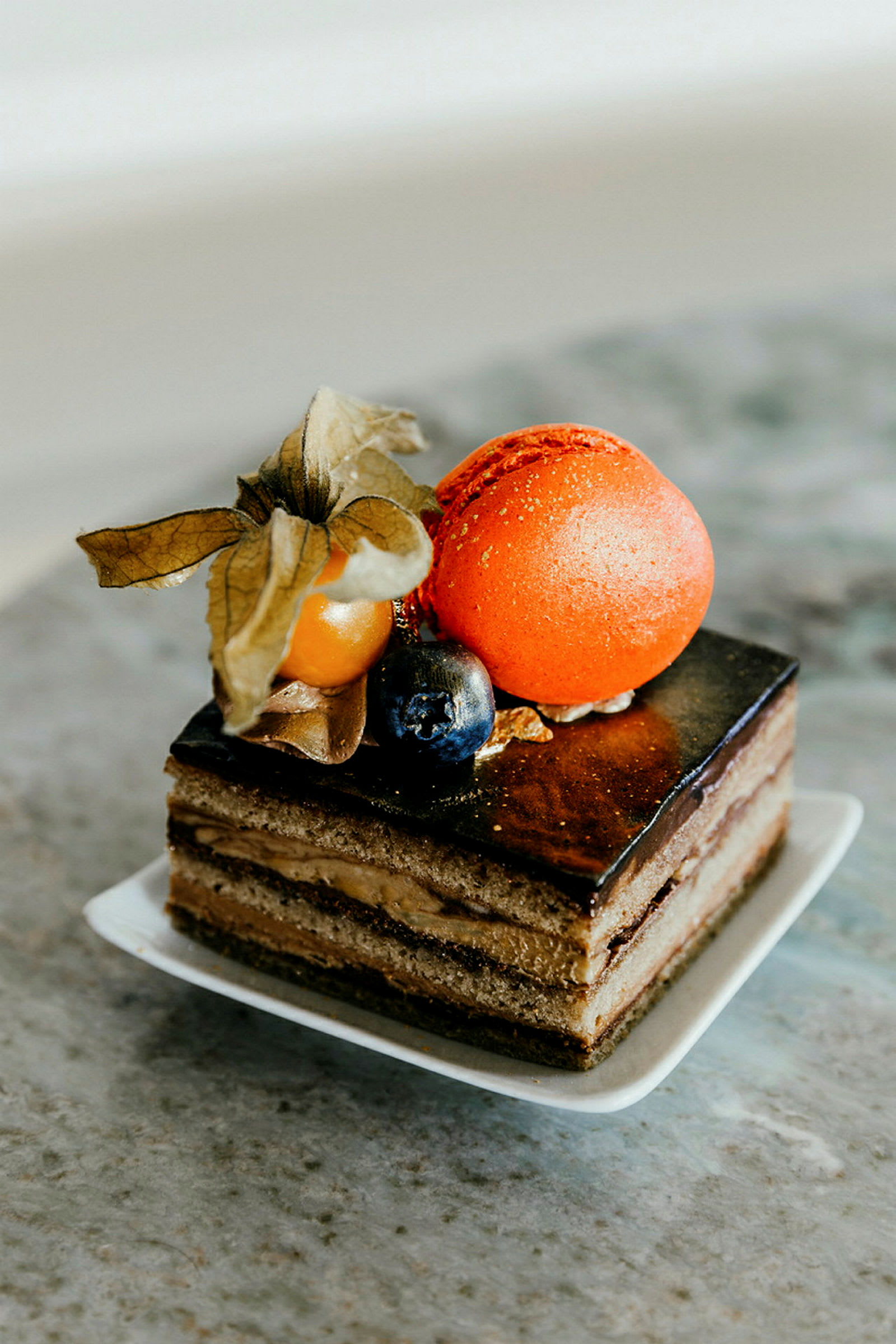 Närbild av en elegant dessert med chokladglasyr, lager av kräm, dekorerad med en orange macaron, en physalis och en blåbär. (AI-genererad text)