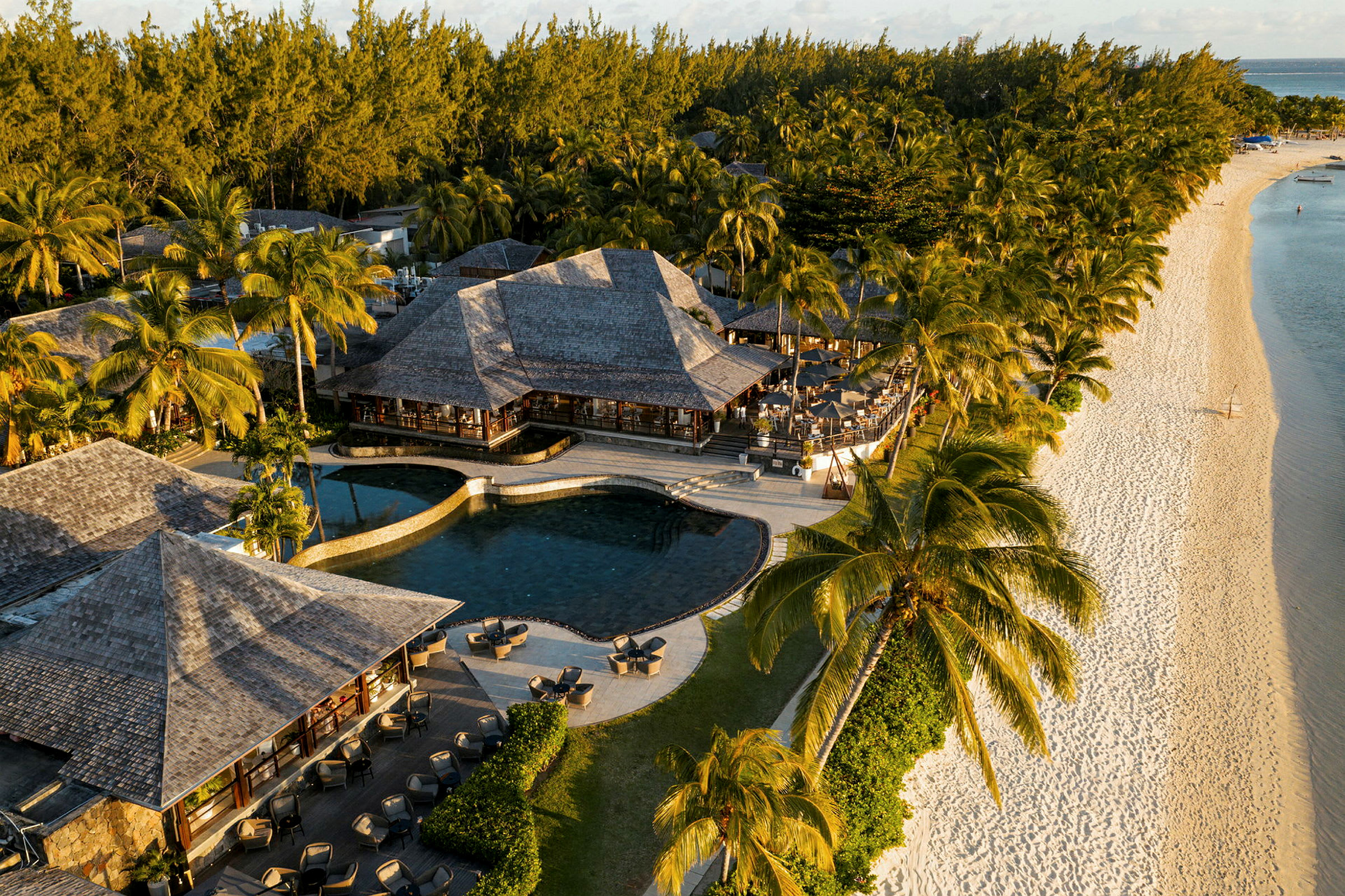 Flygfoto av en resort vid en strand med pooler, byggnader och omgivande palmträd. (AI-genererad text)