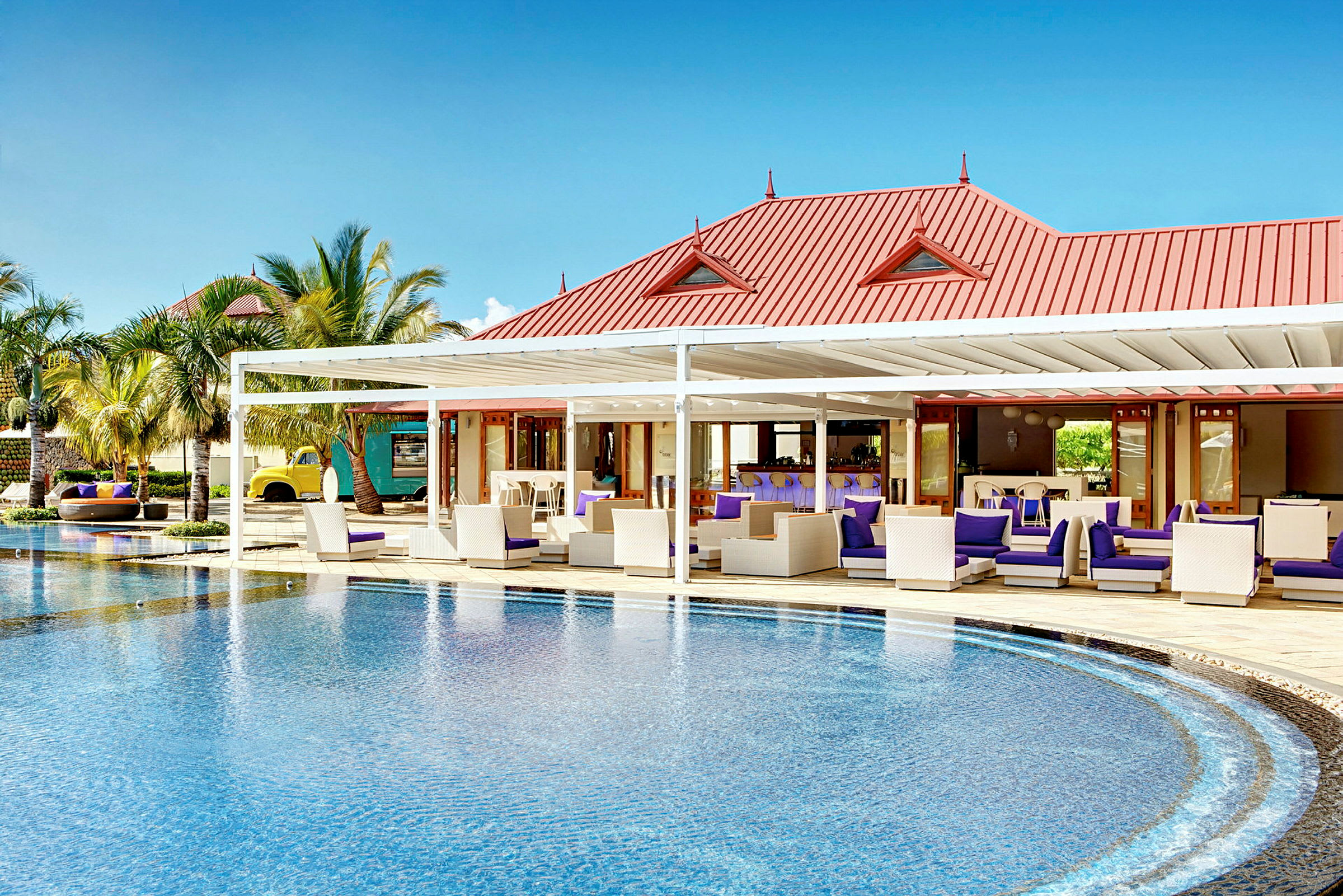 En elegant pool med en loungeområde och en byggnad med rött tak i bakgrunden. (AI-genererad text)