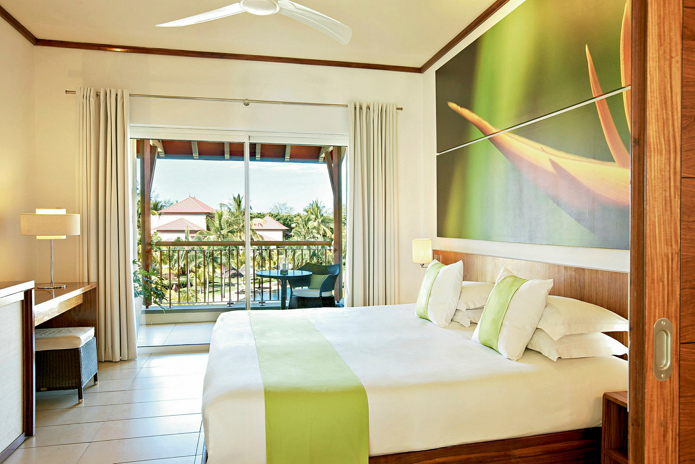 Hotellrum med en dubbelsäng, en stor fönstervägg och en balkong med utsikt över gröna områden och tak av intilliggande byggnader. (AI-genererad text)