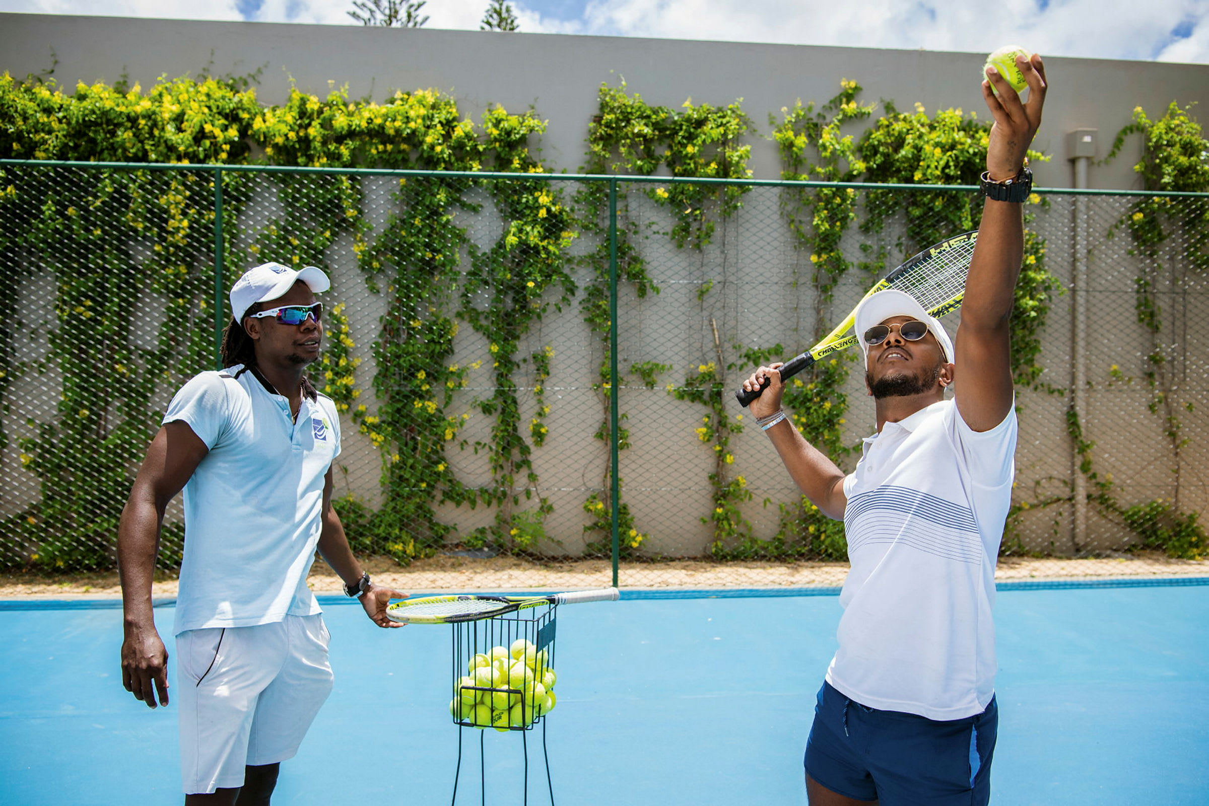 Två personer spelar tennis på en utomhus tennisbana med en behållare fylld med tennisbollar. (AI-genererad text)