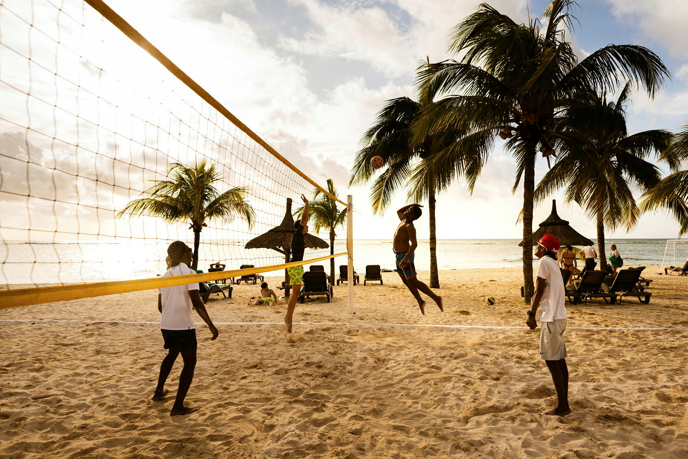 Fyra personer spelar beachvolleyboll på en sandstrand med solstolar och palmer i bakgrunden. (AI-genererad text)