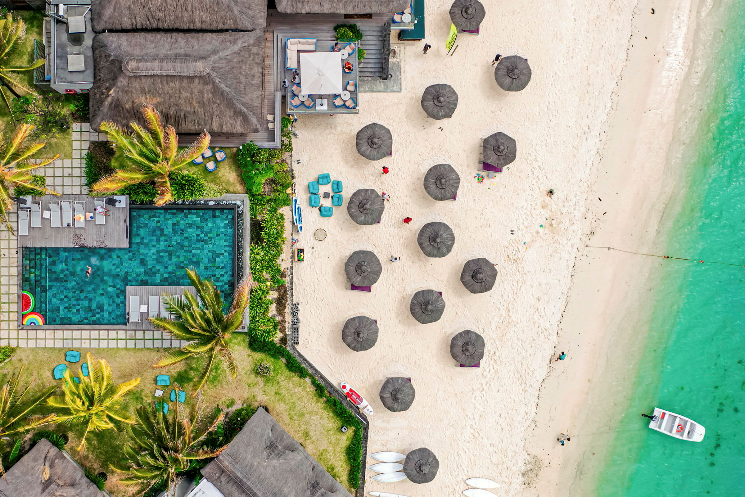 Fågelperspektiv på en strand med parasoller, grön pool, frodig vegetation och turkost hav. (AI-genererad text)
