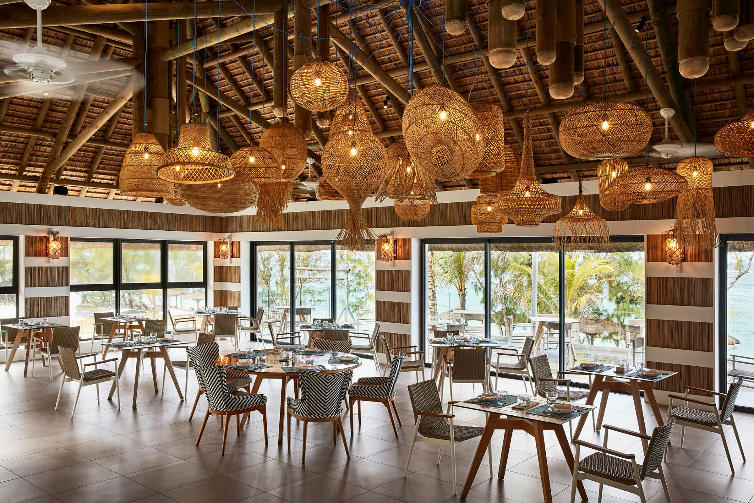 En restaurang med bord och stolar, stora fönster med utsikt mot tropisk miljö och dekorativa taklampor. (AI-genererad text)