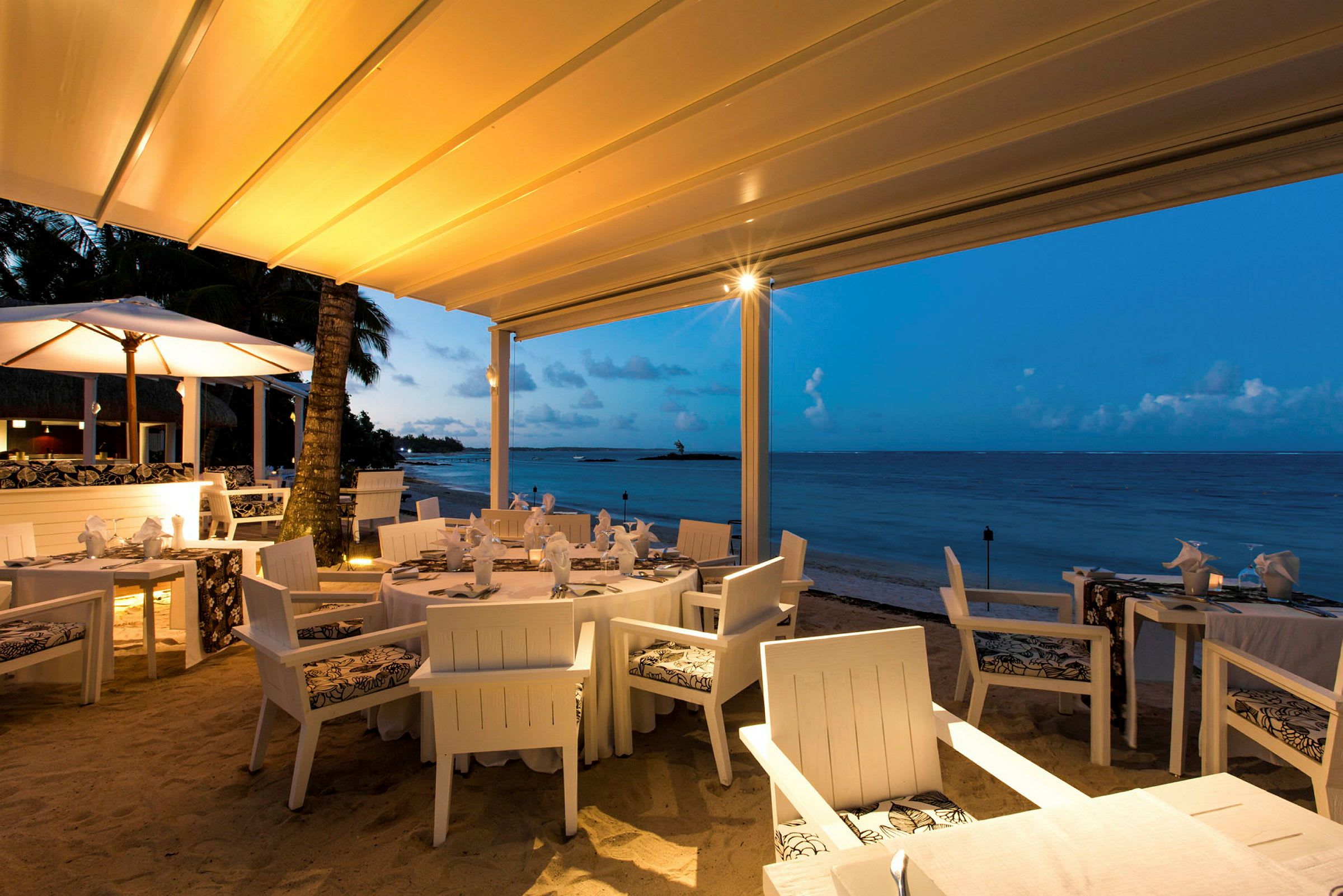 En restaurang med vit möblering är belägen vid stranden under en tältliknande takstruktur. Havet syns i bakgrunden med en klarblå himmel och diskreta moln. (AI-genererad text)