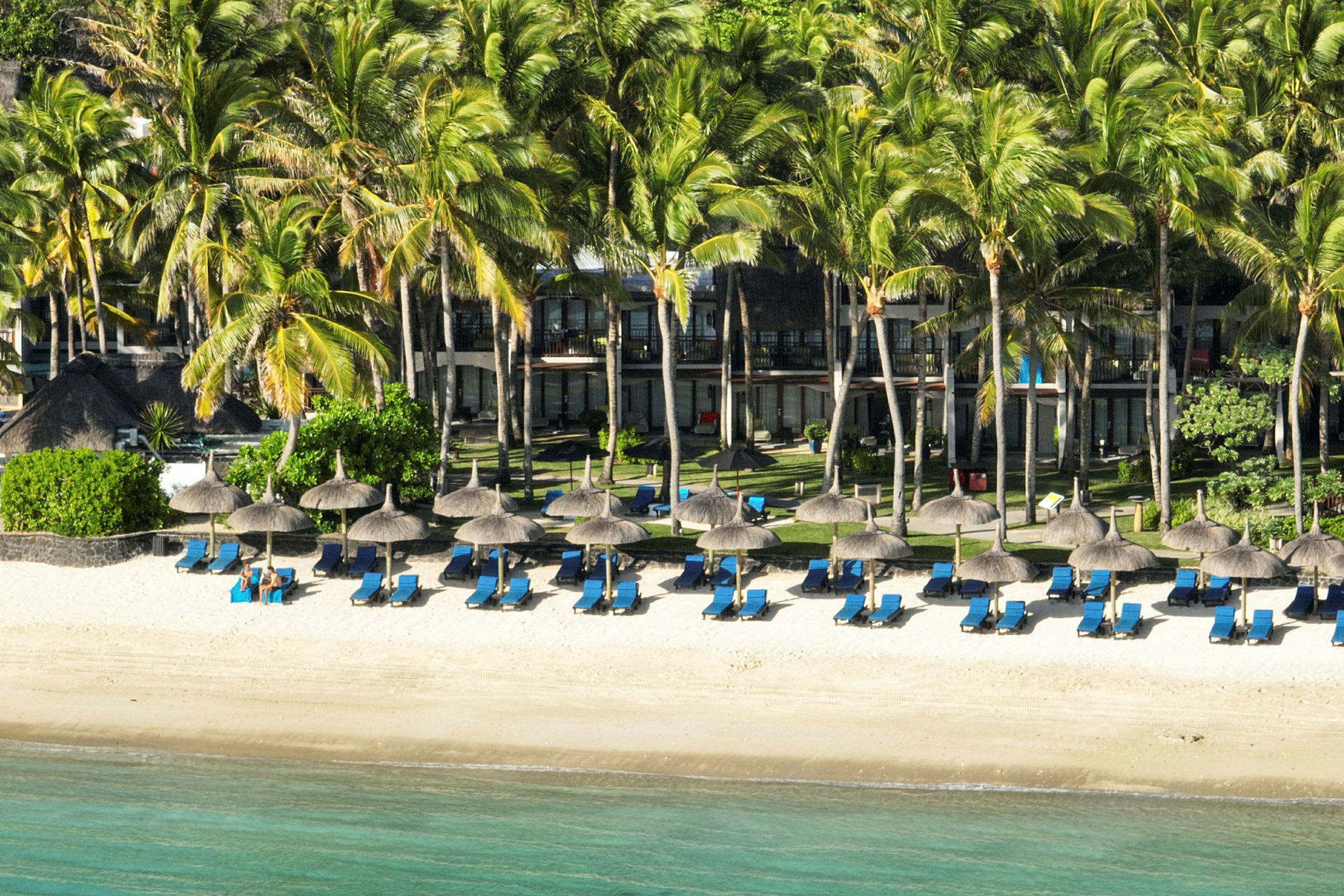 Strand med solstolar och parasoller framför ett hotell omgivet av palmer. (AI-genererad text)