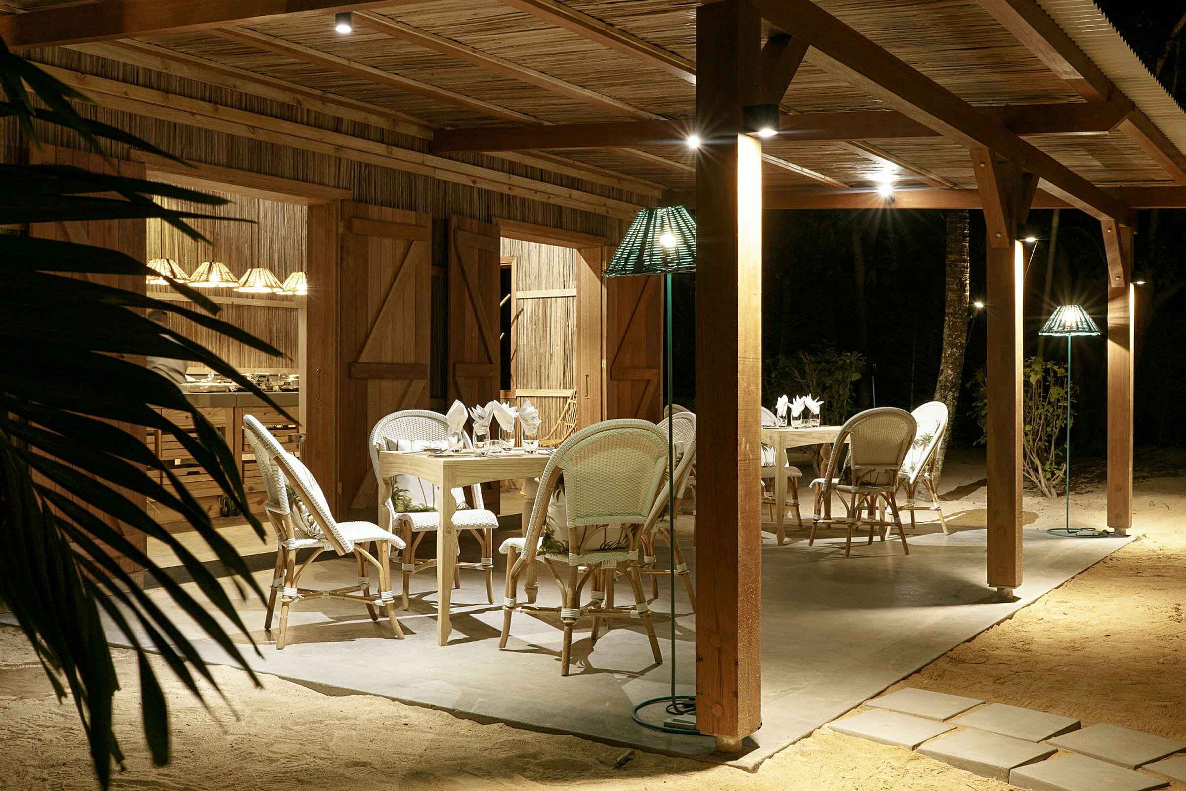 Utomhusområde med bord och stolar under träpergola, kvällsbelysning och sandmark. (AI-genererad text)