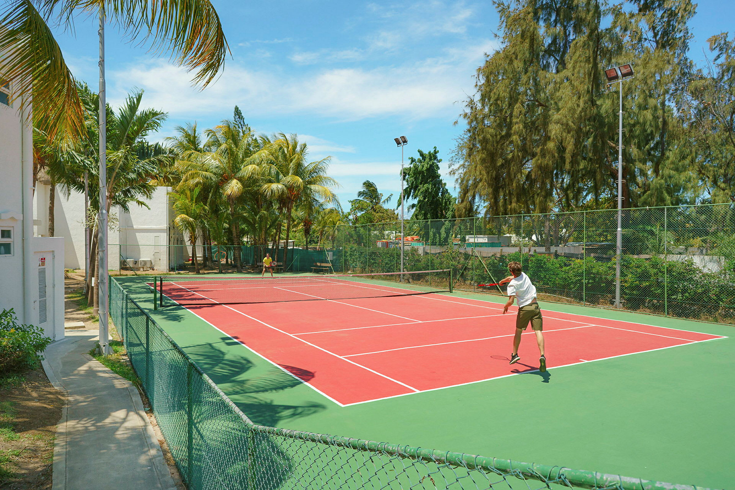 Tennisbana utomhus med två spelare nära ett staket och palmträd. (AI-genererad text)