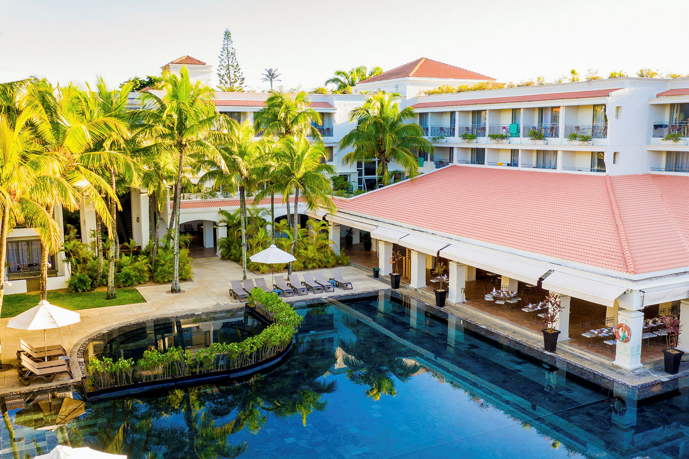 Ett hotellområde med en pool omgiven av solstolar och parasoller, samt palmer och en byggnad med röda tak. (AI-genererad text)
