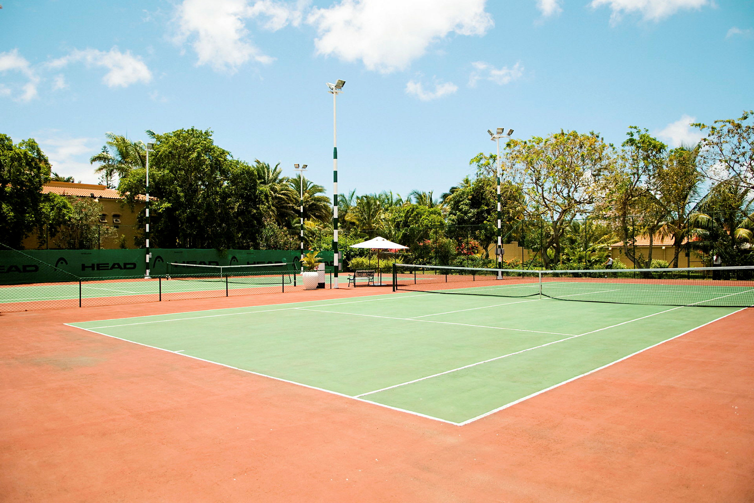 En tennisbana med grönt spelområde och röda omgivande ytor, omgiven av träd och byggnaden. (AI-genererad text)