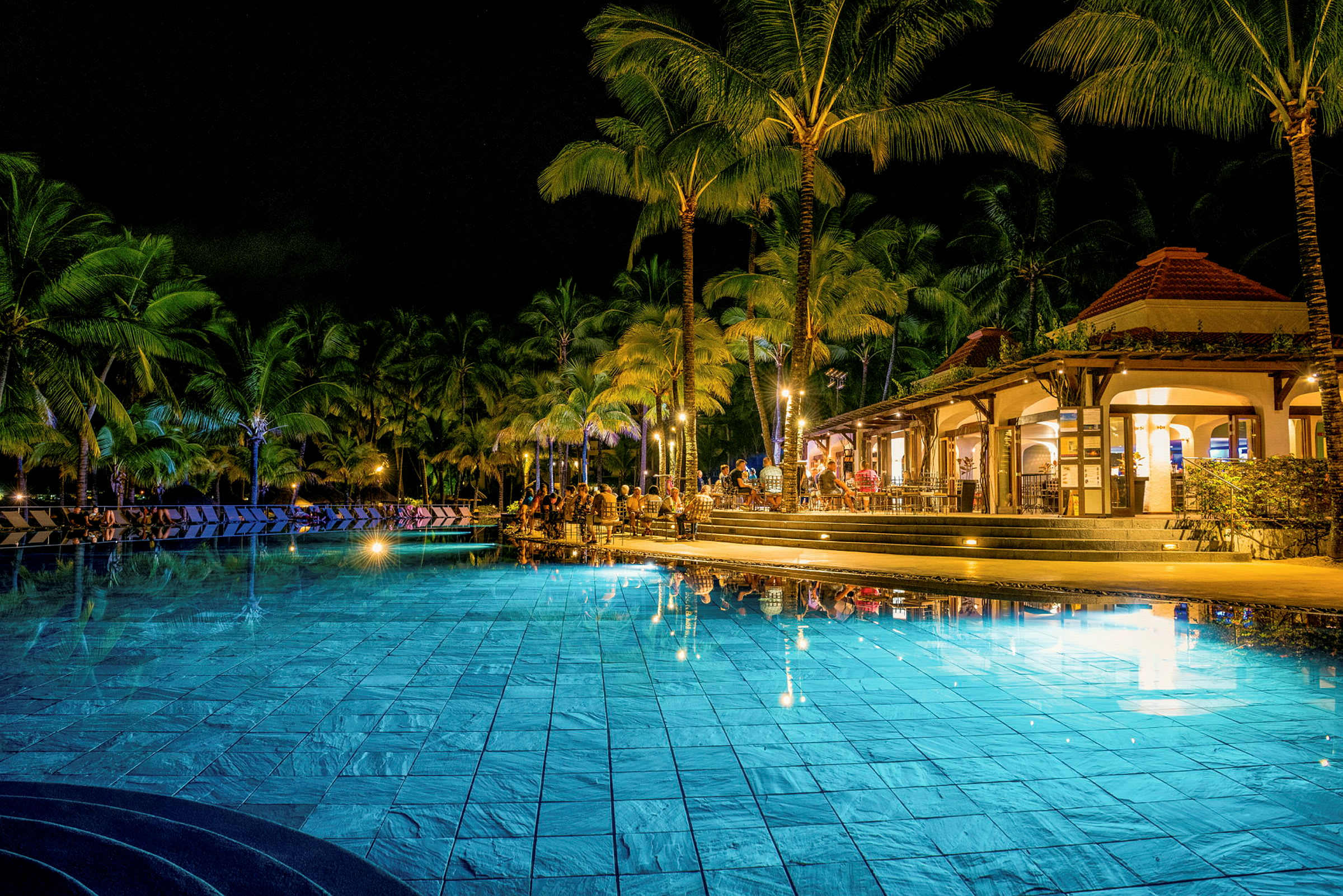 Kvällsbild av en upplyst hotellpool omgiven av palmträd och en restaurang. (AI-genererad text)