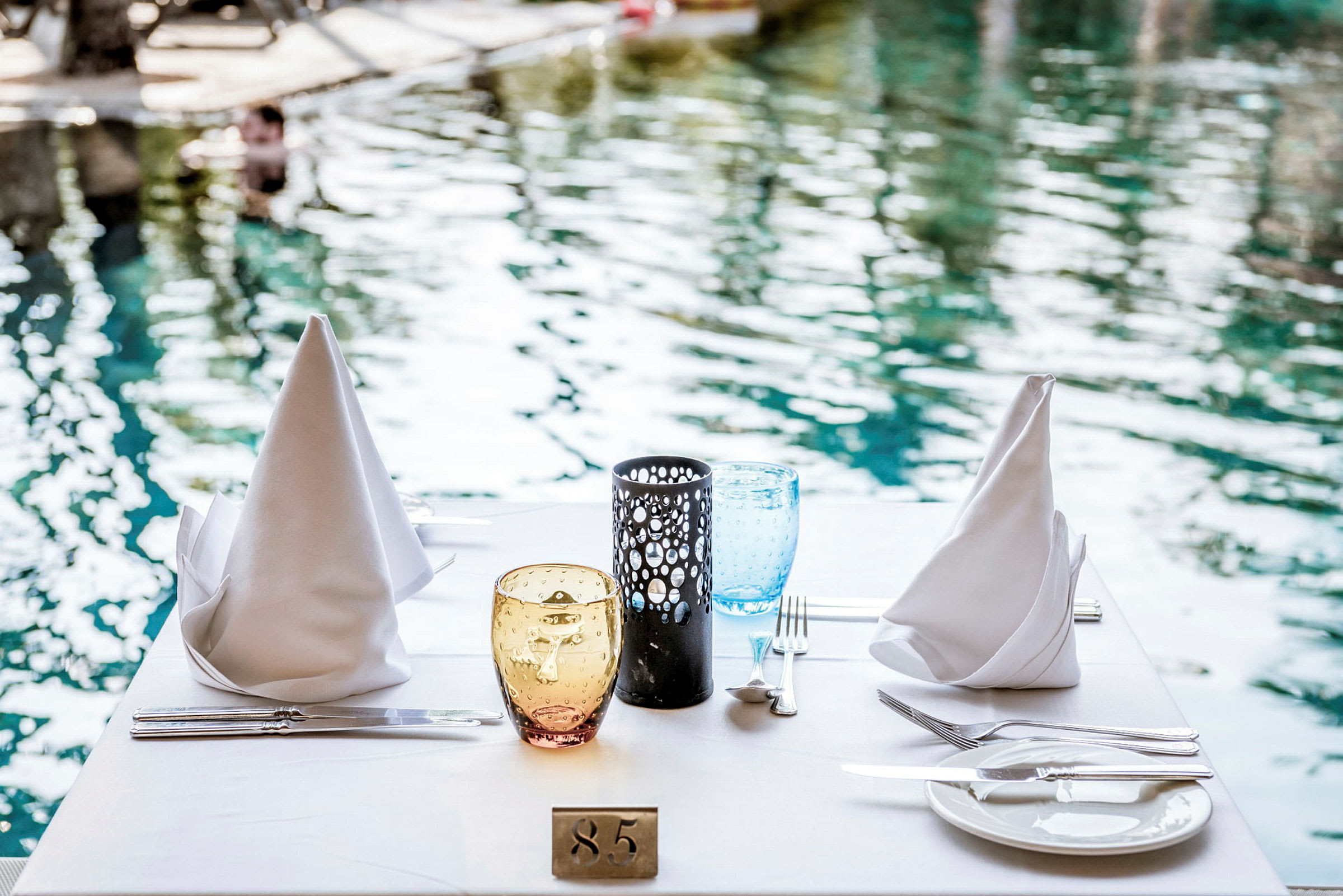 Ett dukat bord med vita servetter, glas och porslin vid sidan av en pool. (AI-genererad text)