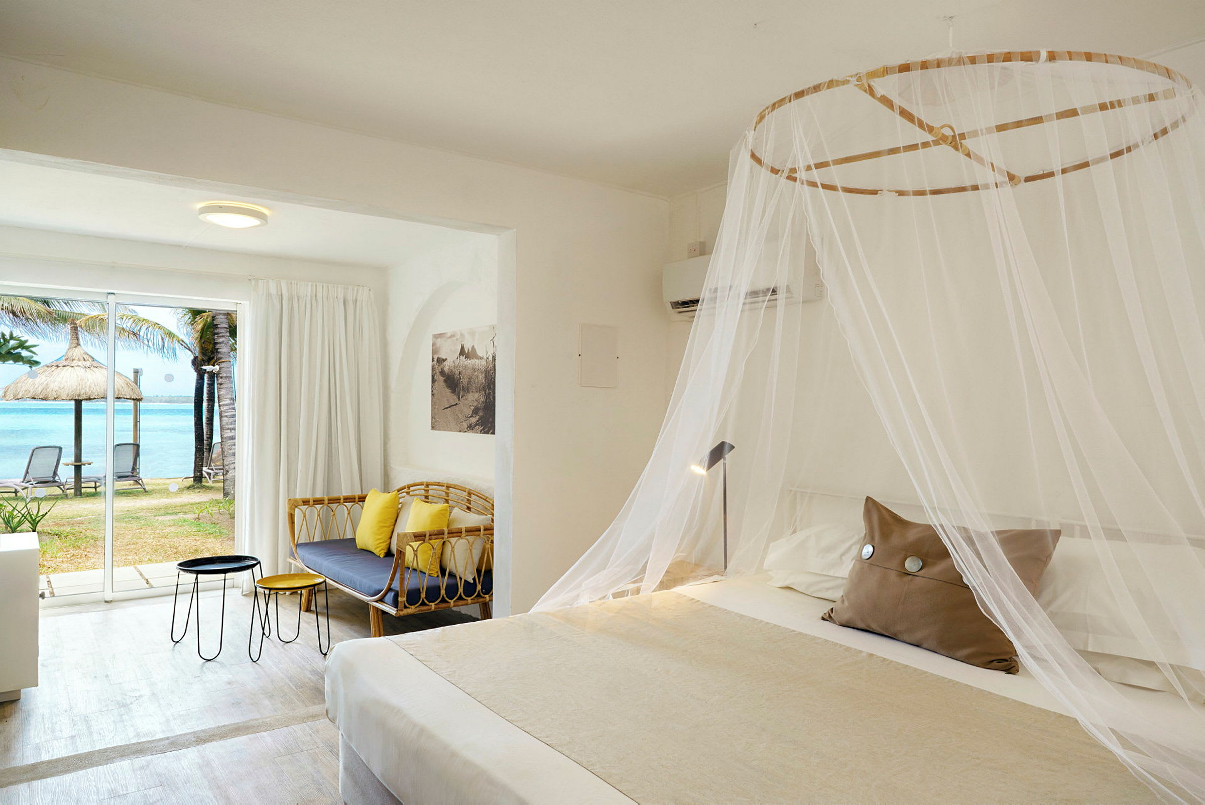 Hotellrum med en stor säng täckt med ett myggnät, en soffa, bord och ett stort fönster med utsikt över stranden och havet. (AI-genererad text)