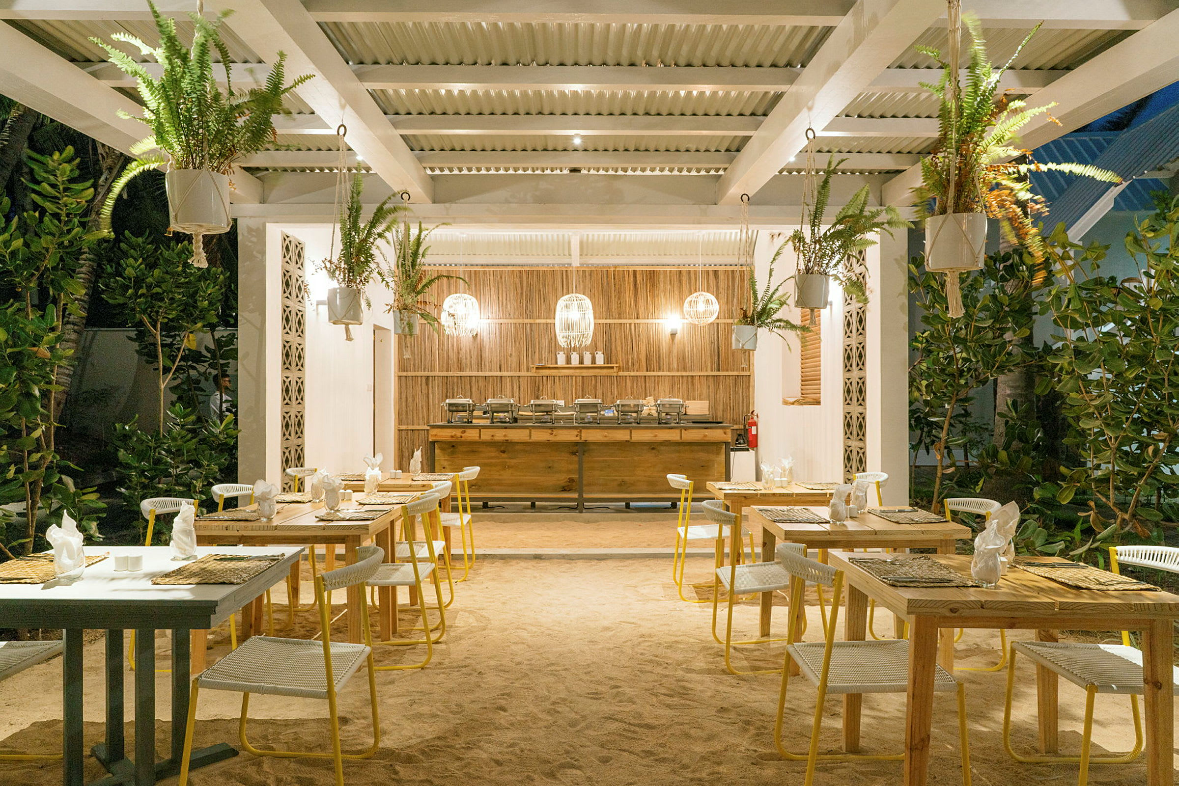 Utomhusrestaurang med bord och stolar på sandgolv, omgiven av grönska. (AI-genererad text)