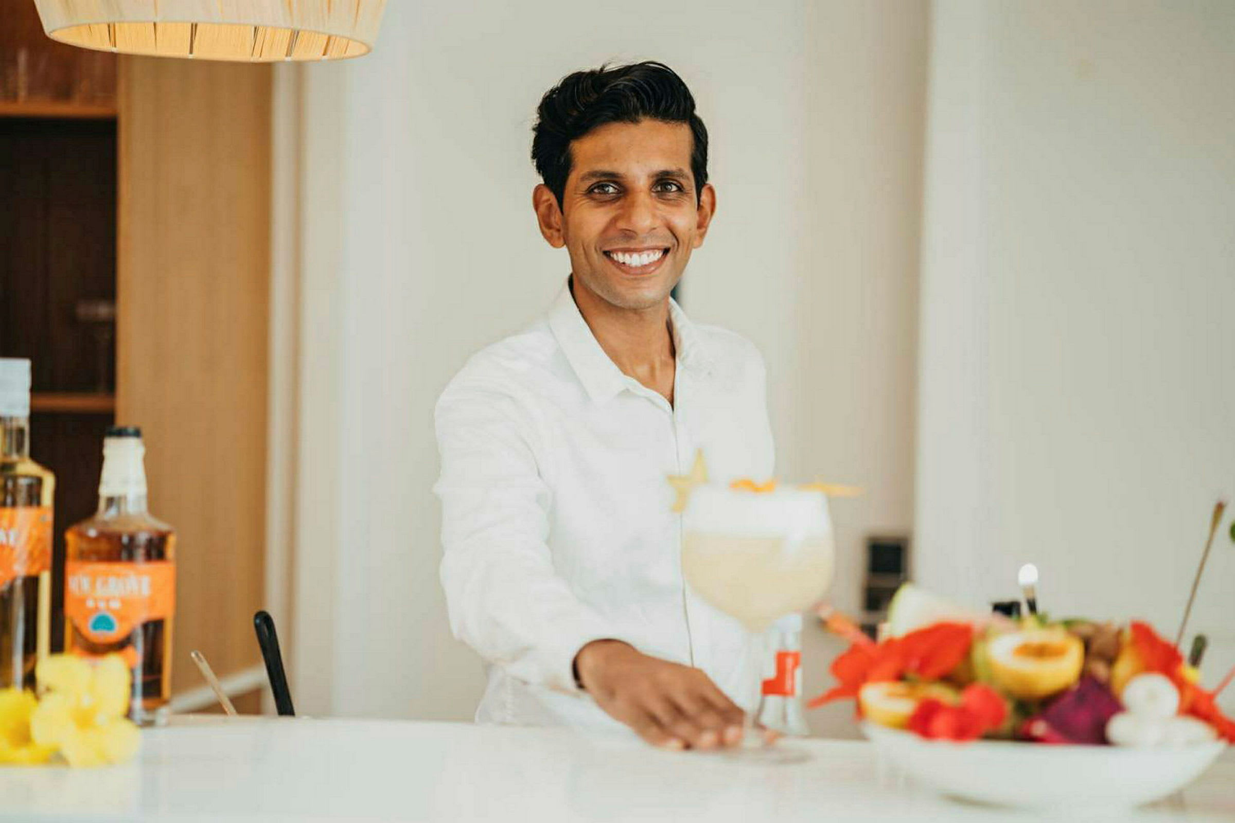 Person i vit skjorta vid en bardisk med en stor cocktail och en skål färsk frukt. (AI-genererad text)