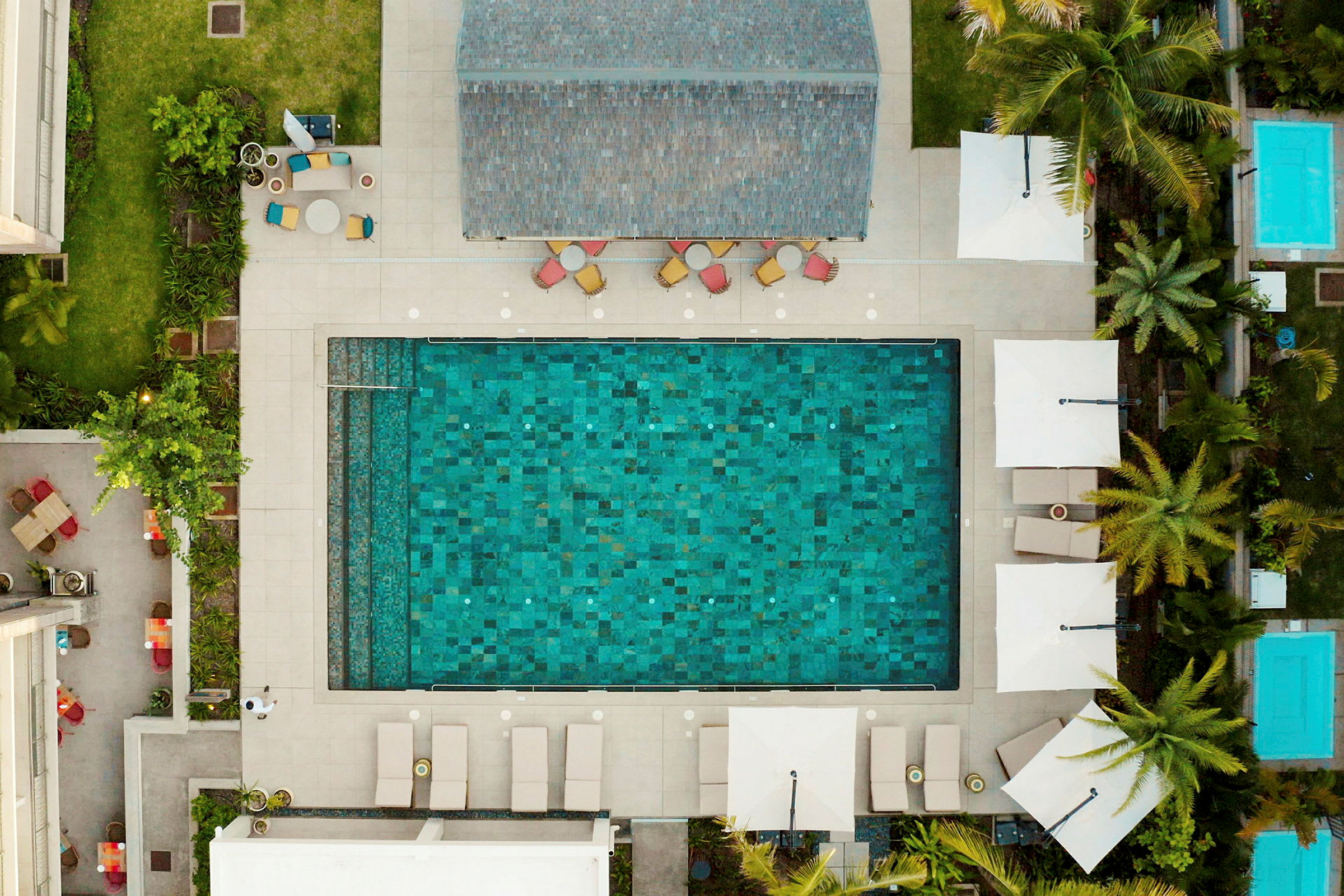 Flygfoto av en rektangulär pool omgivet av solstolar, parasoller och gröna växter. (AI-genererad text)