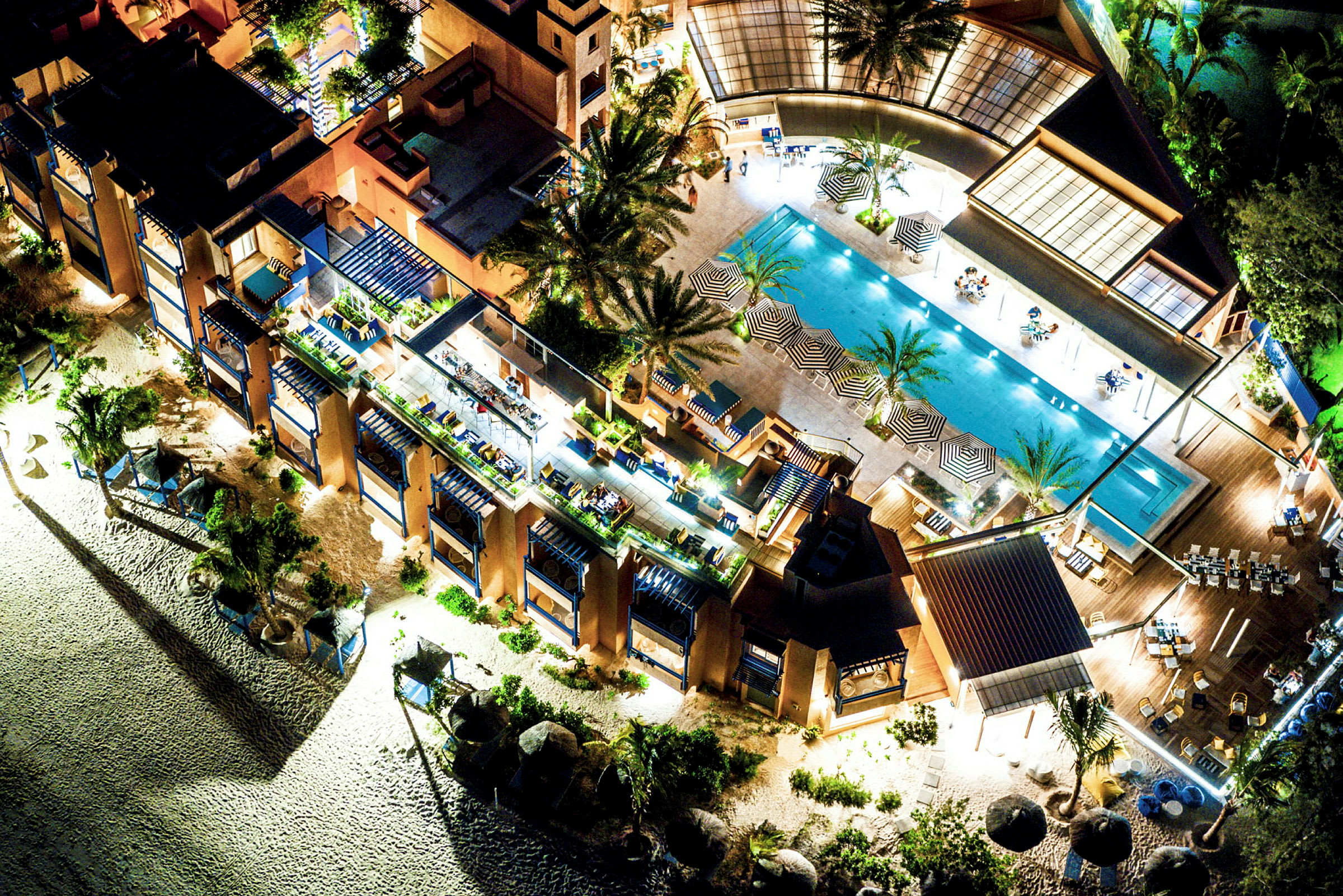 Fågelperspektiv över en resort med pool, solstolar, parasoller och omgivande byggnader. (AI-genererad text)