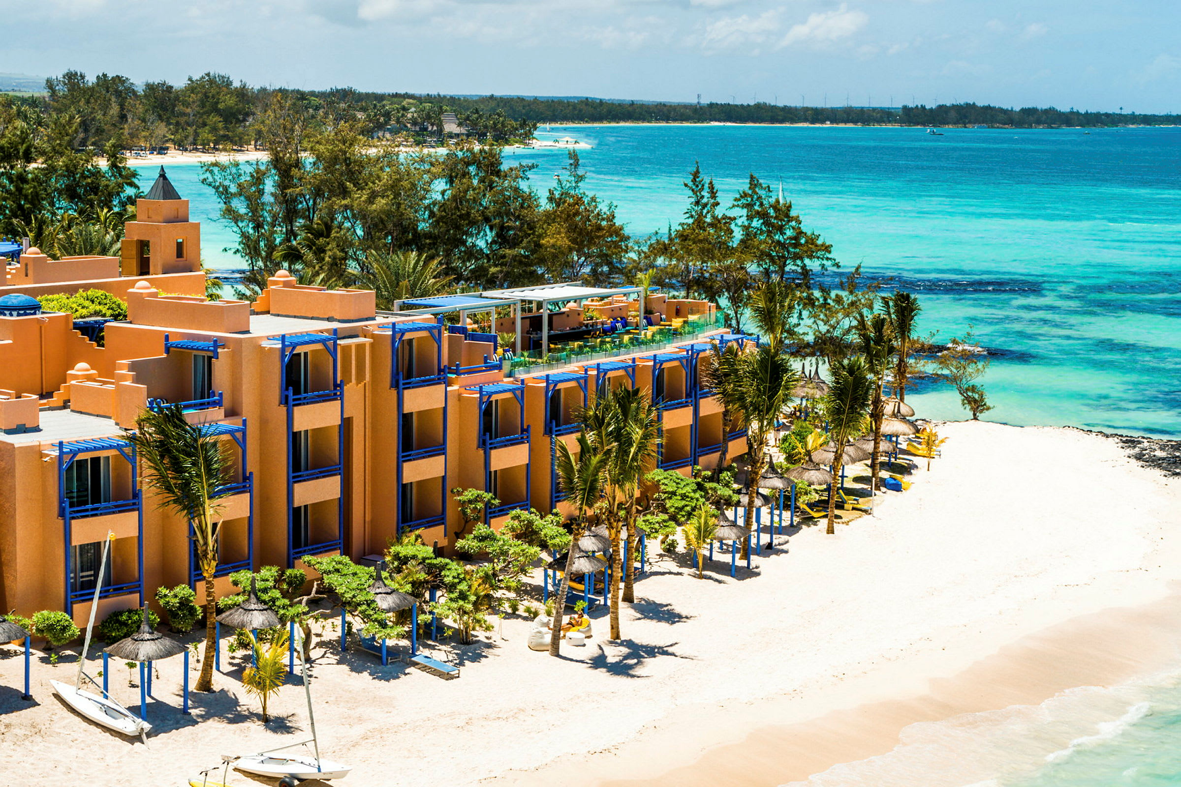 Hotellbyggnad vid en vit sandstrand med blå parasoller och havet i bakgrunden. (AI-genererad text)