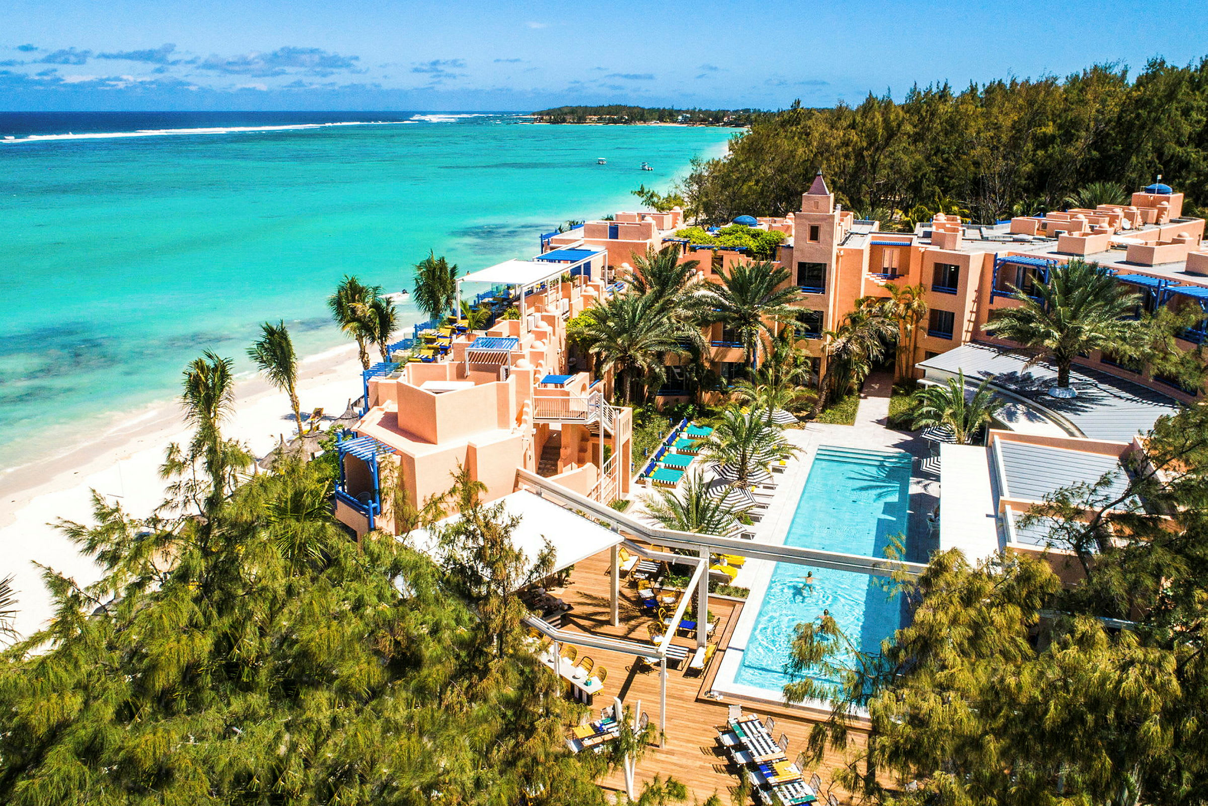 Flygvy över ett hotellkomplex vid en tropisk strand med pool och omgivande grönska. (AI-genererad text)