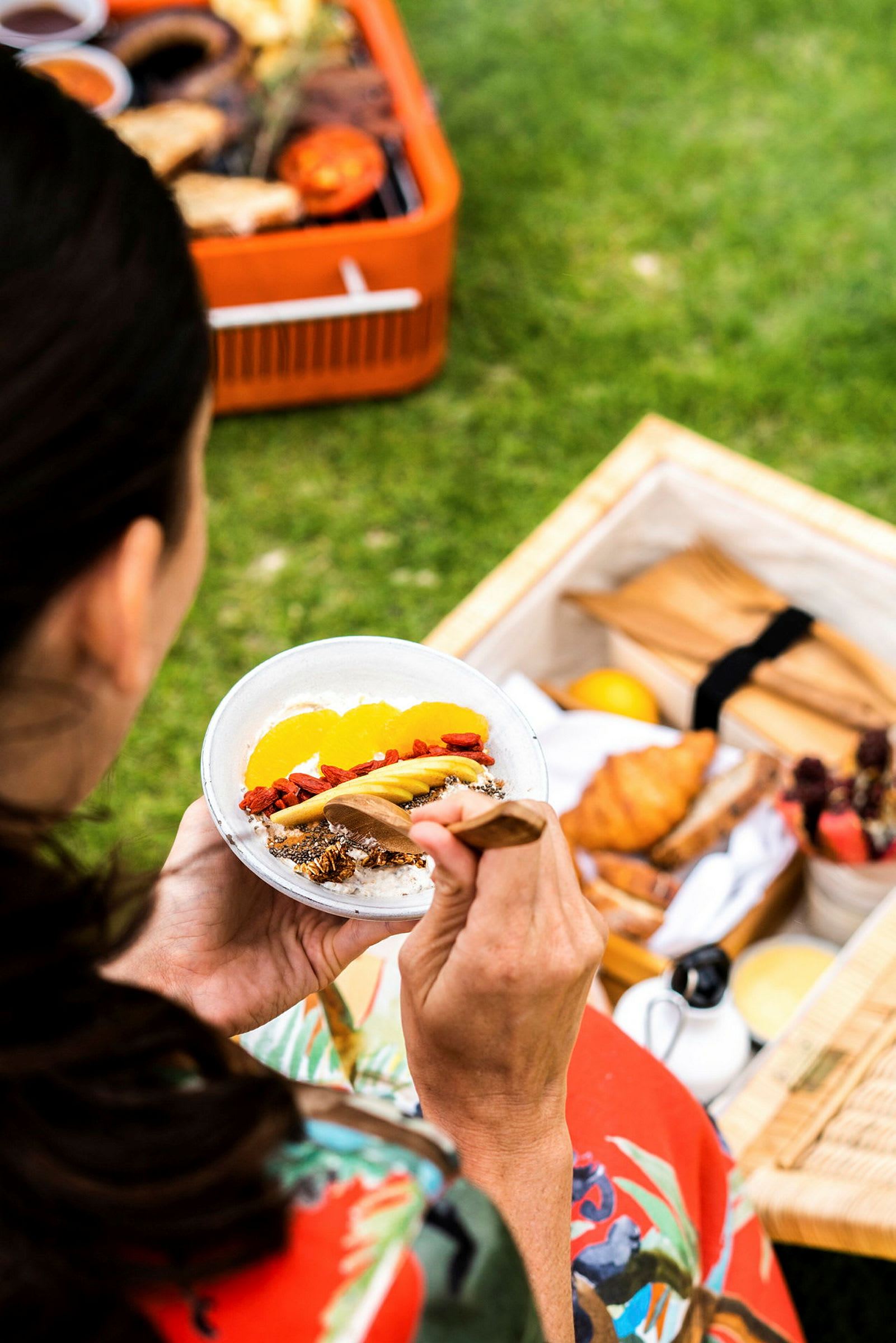 En person håller en tallrik med yoghurt och frukt, omgiven av en picknickuppsättning utomhus. (AI-genererad text)