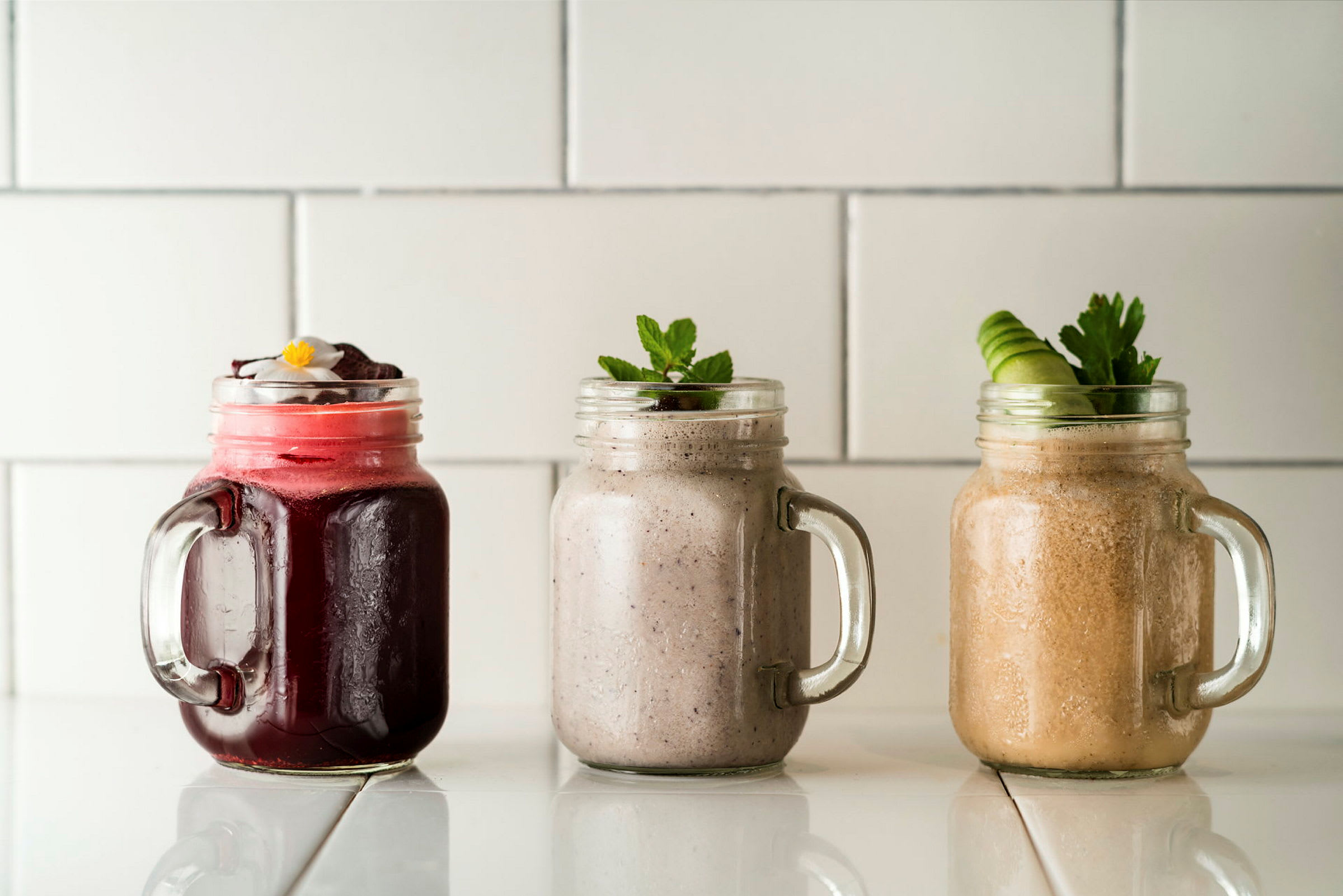 Tre olika smoothies i glasburkar på ett vitt underlag. (AI-genererad text)