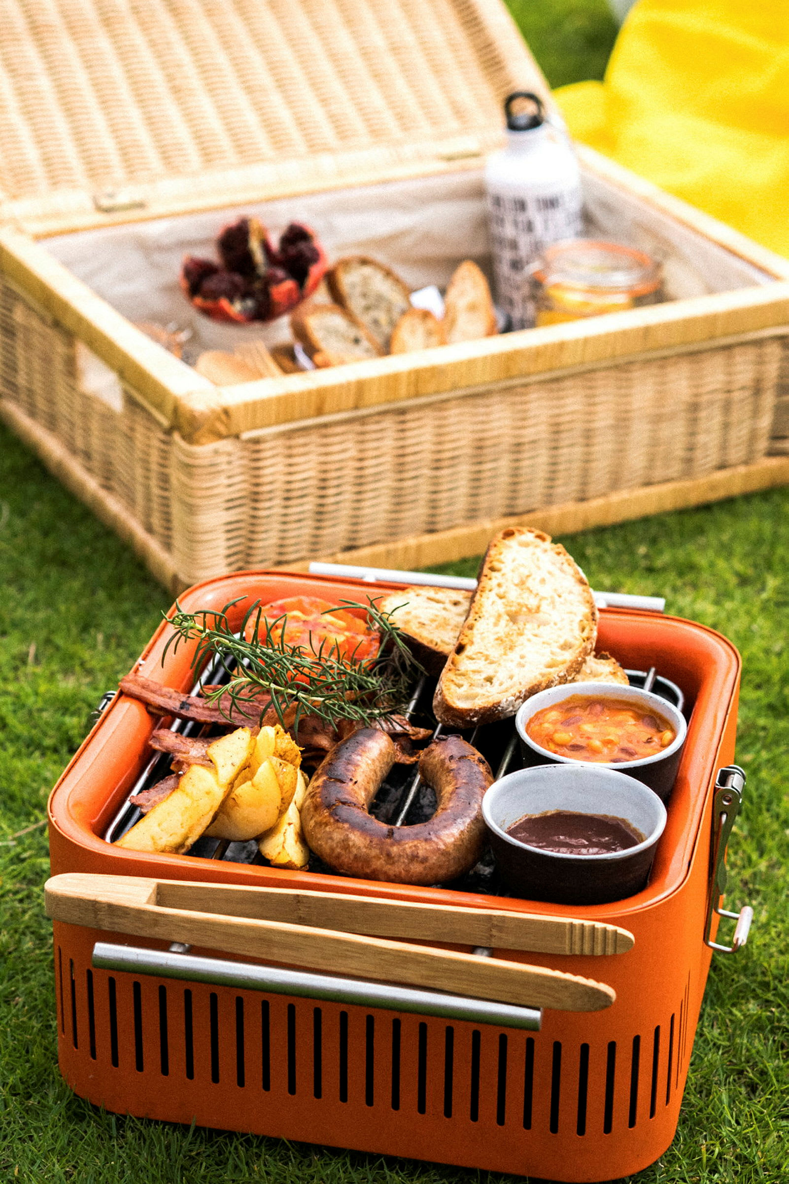 En orange grill med mat, inklusive korv och bröd, står på gräs. I bakgrunden finns en korg med picknicktillbehör. (AI-genererad text)