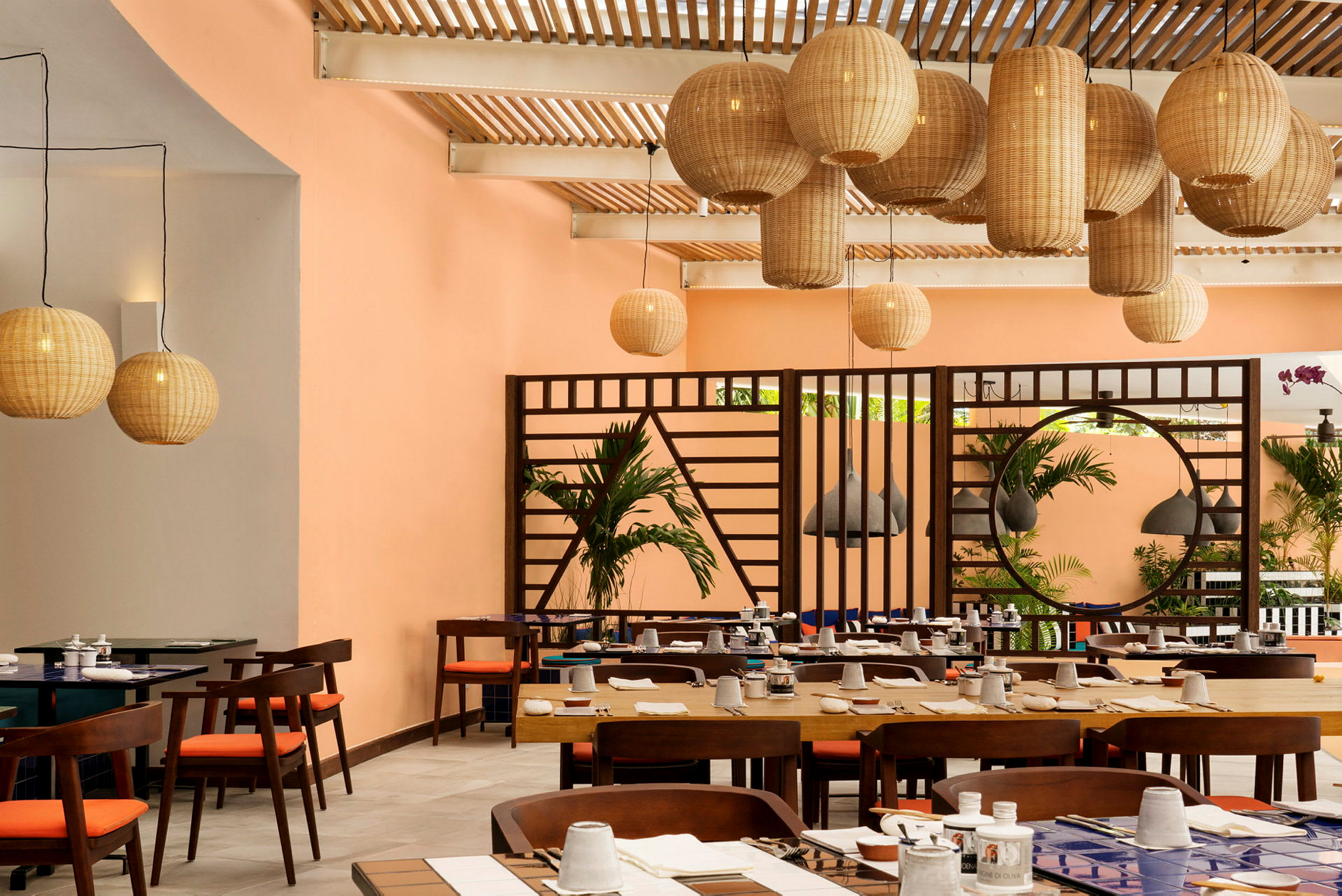 En inomhus restaurang med bordsdukar och hängande bastlampor. Bakgrunden visar orange väggar och grönska. (AI-genererad text)