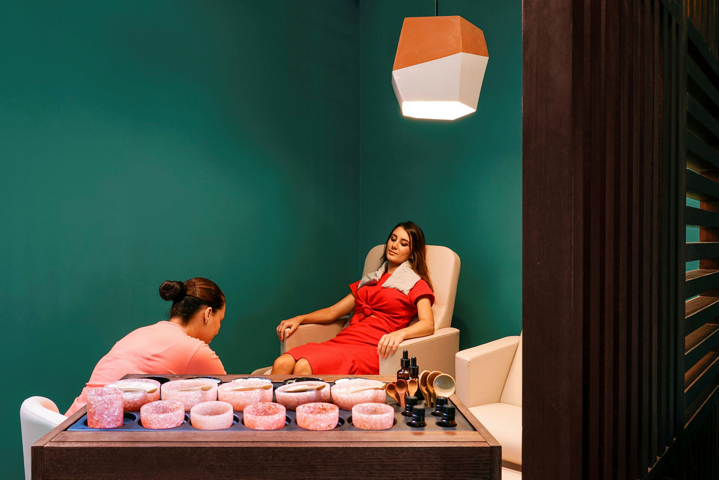 Ett spa-rum med en person som får en behandling, medan en annan person assisterar. I förgrunden finns en träig disk med rosa saltstenar och skönhetsprodukter. (AI-genererad text)