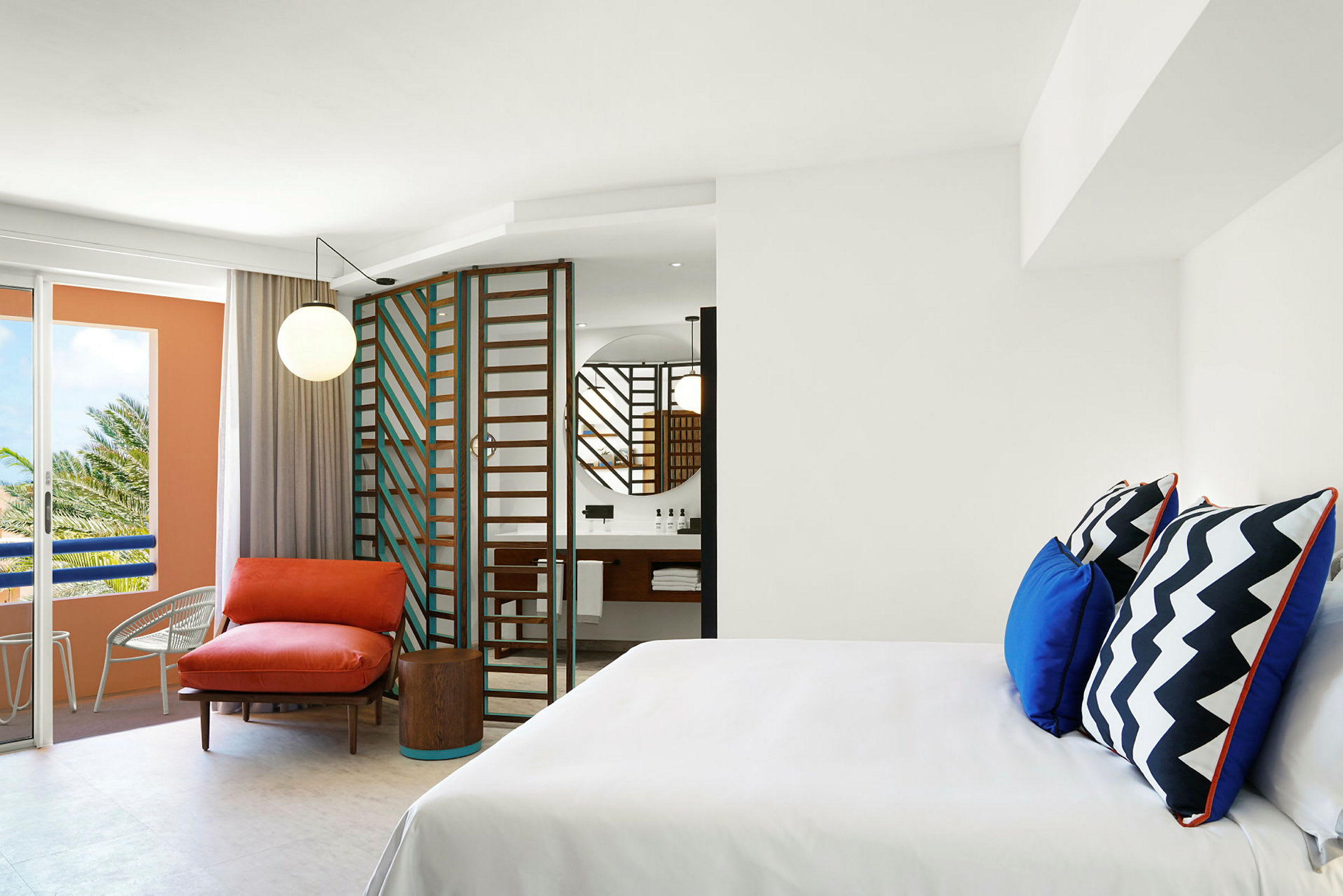 Ett modernt hotellrum med vita väggar, en färgglad fåtölj och kuddar samt ett badrum med spegel och en glasdörr. (AI-genererad text)