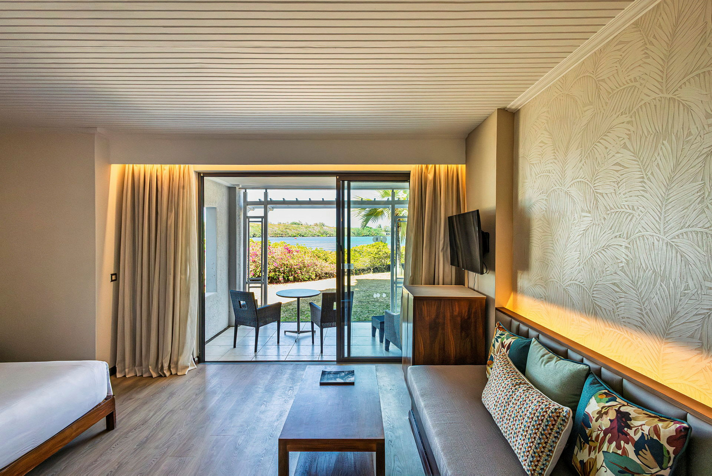Ett hotellrum med modern inredning, en soffa med färgglada kuddar, ett bord och stolar samt en balkong med utsikt över en trädgård och vatten. (AI-genererad text)