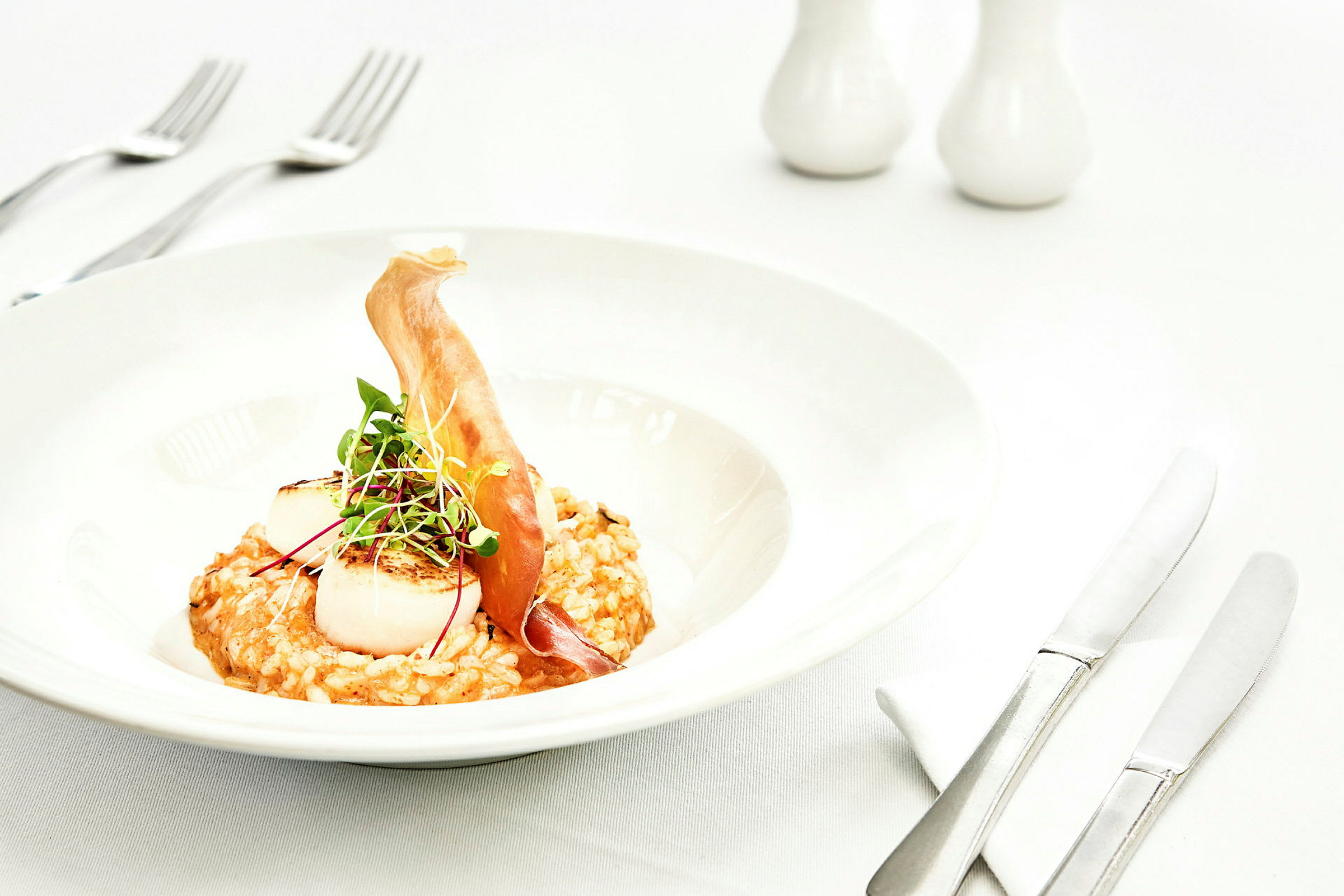 Tallrik med risotto, grillade pilgrimsmusslor och en dekoration av mikrogrönt samt tunt skivade råvaror. (AI-genererad text)