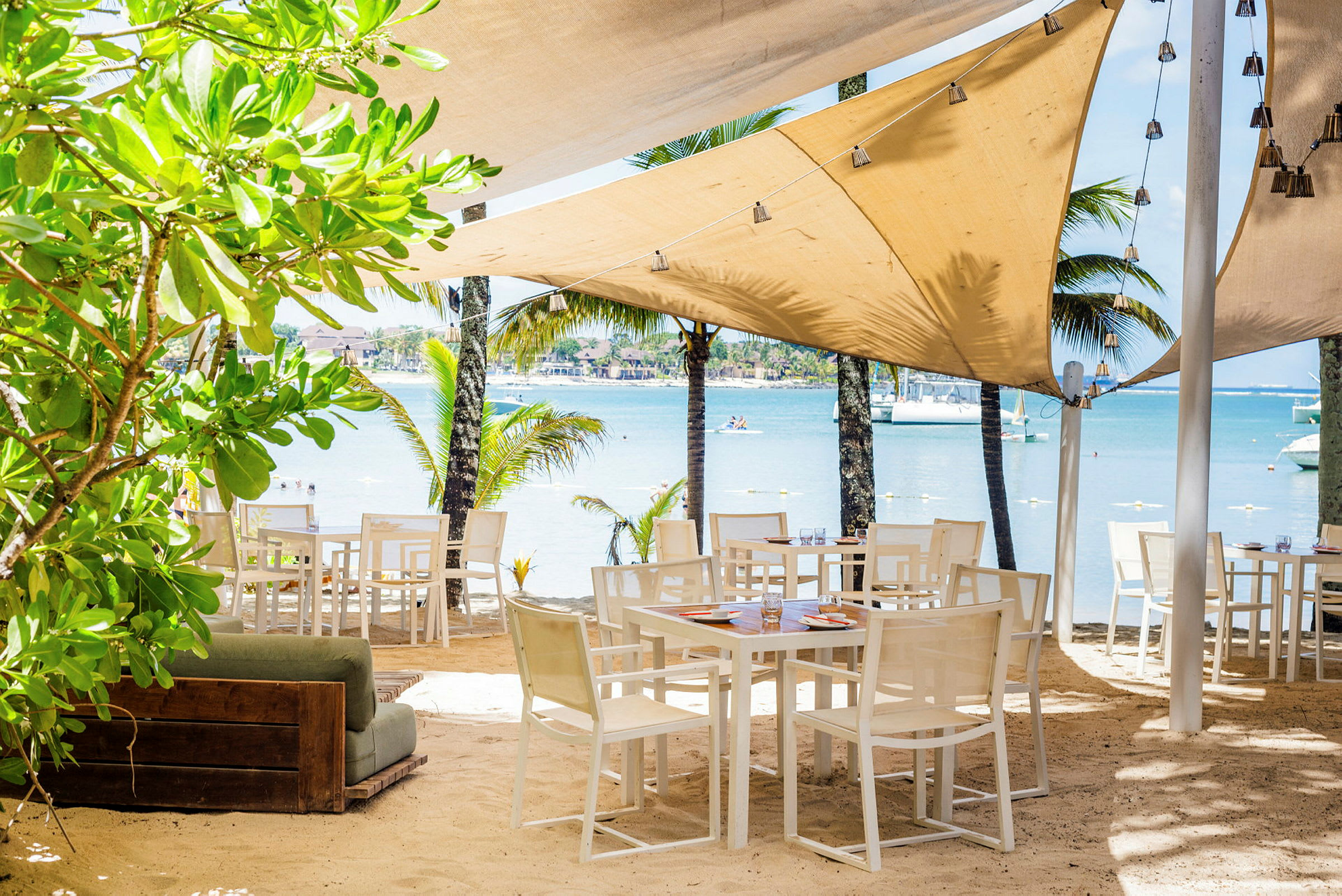 Utomhusrestaurang med bord och stolar på sandstrand, omgiven av grönska och palmer med utsikt över havet. (AI-genererad text)
