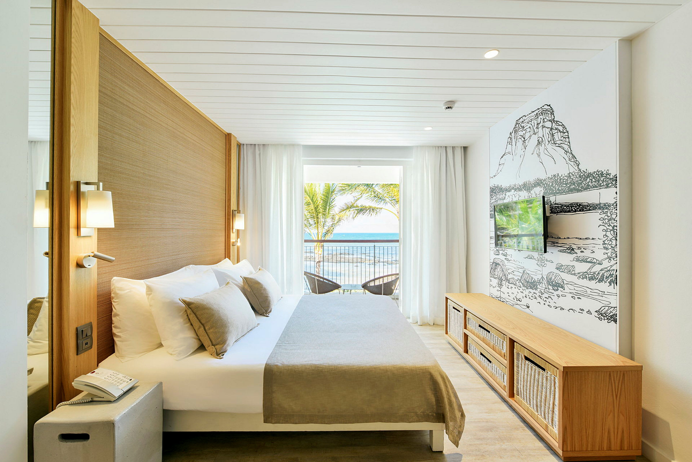 Ett ljust hotellrum med en dubbelsäng, väggmonterad TV och stora fönster som vätter ut mot havet. (AI-genererad text)