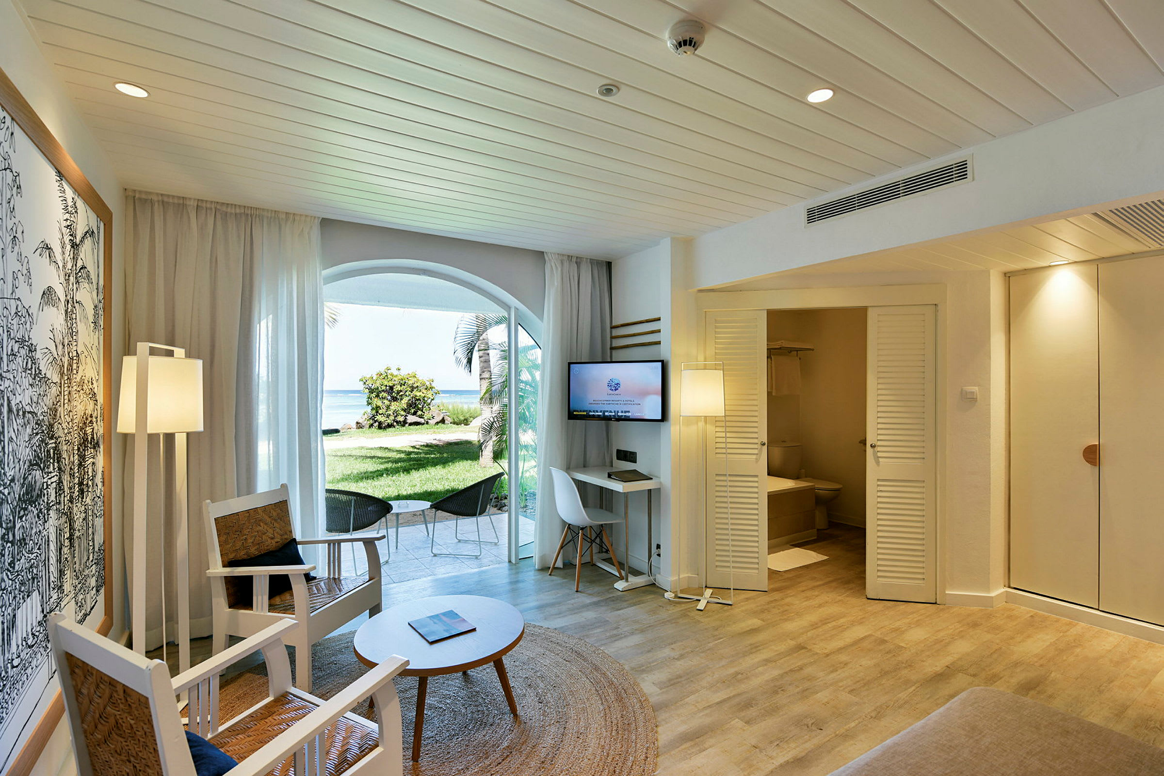 Ett hotellrum med sittmöbler, ett bord och en TV. En dörr leder till ett badrum, och det finns fönster som vetter mot en grön trädgård och stranden. (AI-genererad text)