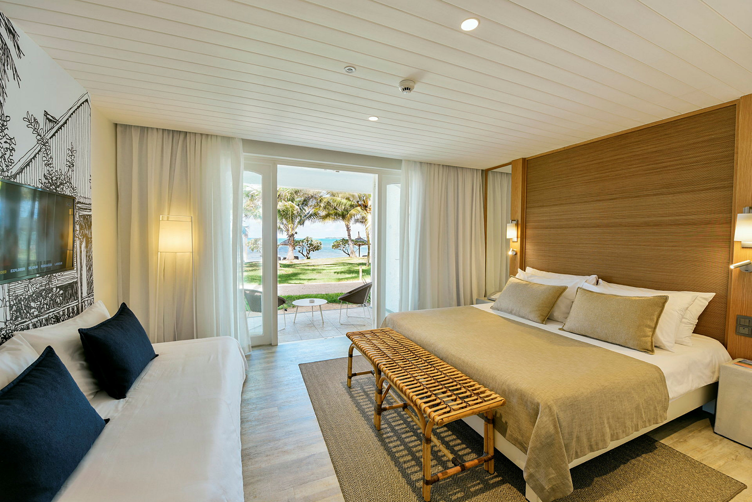 En hotellrum med en dubbelsäng, soffa, och en balkong med utsikt över tropisk natur samt havet. (AI-genererad text)